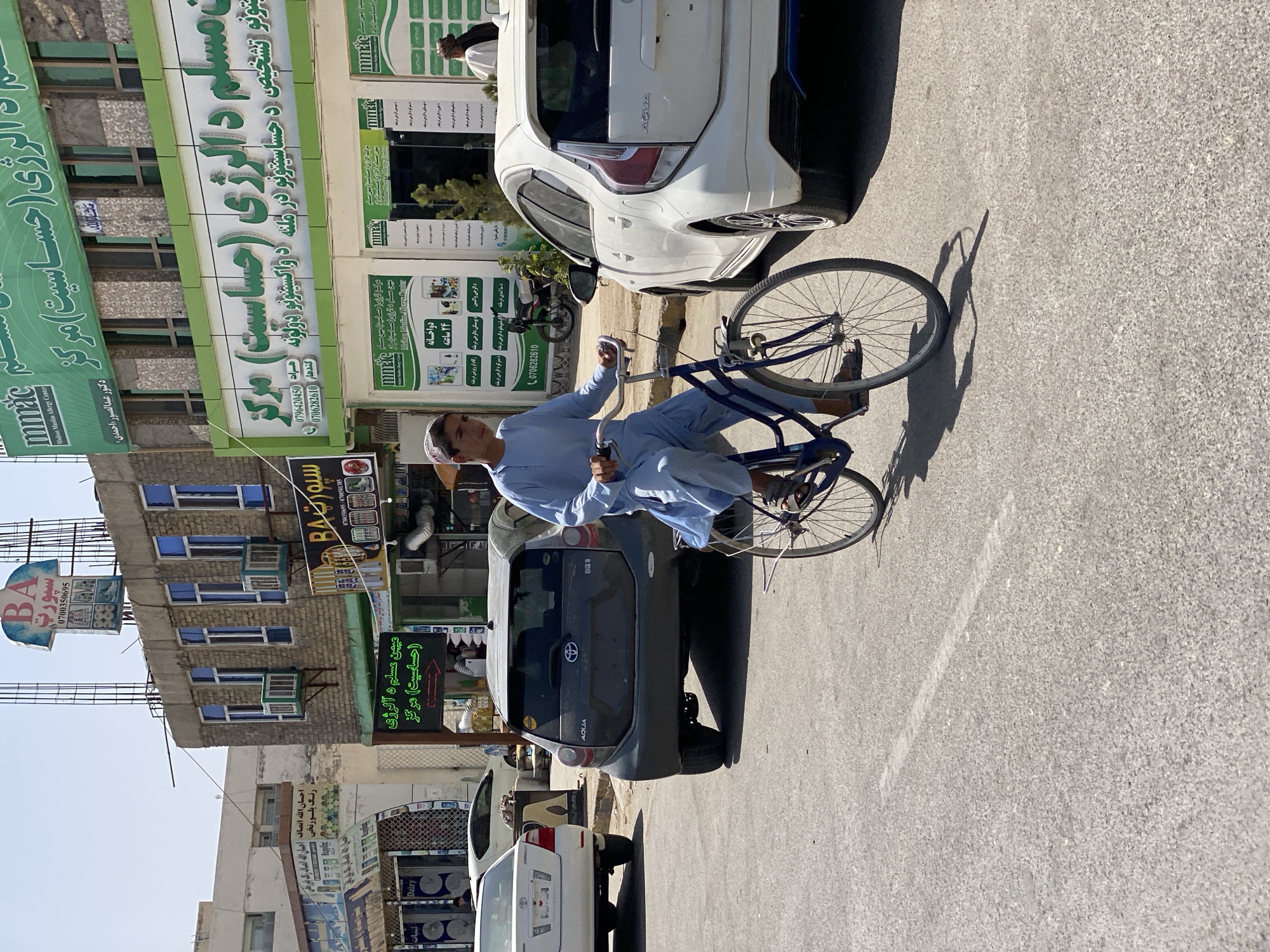 In Kandahar, Afghanistan, a young man wearing a light blue shalwar kameez and a white pakol is riding a bicycle down a street. He is positioned in the foreground, moving from left to right. The bicycle is dark blue with chrome handlebars and pedals. Behind him, a dark grey Toyota Aqua is parked on the right side of the street. To the left of the Aqua, a white Toyota sedan is also parked. The street is paved and appears to be a commercial area with buildings lining the right side. One building has a green sign with white Arabic text and a logo that includes the letters "mMAC". Another building features a sign with a blue and white logo shaped like a t-shirt, labeled "BA Sport" and a phone number. The buildings are multi-story with visible windows and air conditioning units. The sky is clear and blue, indicating a sunny day. The environment appears to be a typical urban street scene in Afghanistan.