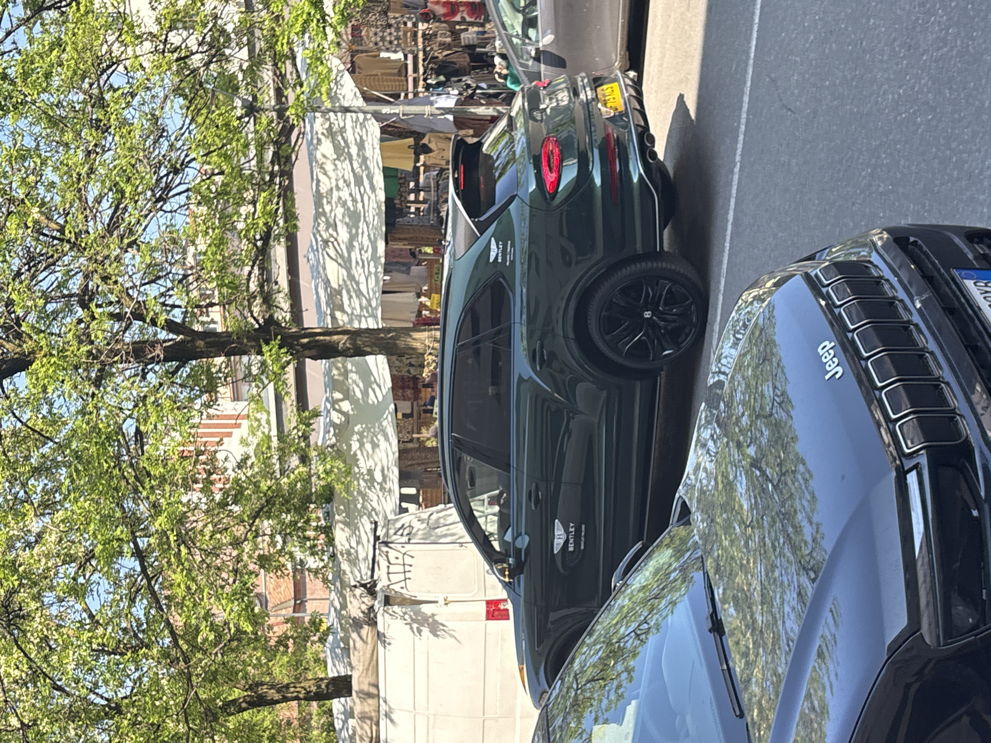 The image presents a sunlit street scene, likely in Milan, Italy, featuring parked luxury vehicles alongside an outdoor market. The perspective is tilted, giving the impression of a clockwise rotation.
In the immediate foreground, the front-right portion of a dark-colored Jeep Grand Cherokee is visible. Its glossy hood reflects the bright green leaves of trees and a clear blue sky, with the "Jeep" logo clearly embossed on its surface.
Behind the Jeep, in the midground, is a dark green Bentley Bentayga SUV. Its side profile prominently displays "BENTLEY" and "BENTAYGA" badging. The vehicle sports distinctive black wheels with the Bentley "B" emblem. The rear of the Bentayga features circular red taillights and quad exhaust pipes. A yellow European license plate is partially visible, showing "PP 13 YLS".
To the left and in the background, numerous trees with vibrant green foliage indicate late spring or early summer. Beyond the trees and cars, an outdoor market is evident, comprising white tented stalls displaying various goods, possibly textiles or clothing. A white commercial van is partially visible among the market structures. A person wearing a green top can be faintly seen near one of the market stalls.
The scene is brightly illuminated by direct sunlight, casting distinct shadows from the trees onto the market tents and from the cars onto the asphalt, suggesting a clear day, likely late morning or afternoon. The combination of high-end vehicles and an active street market points to an urban setting characterized by both commerce and a certain level of affluence.