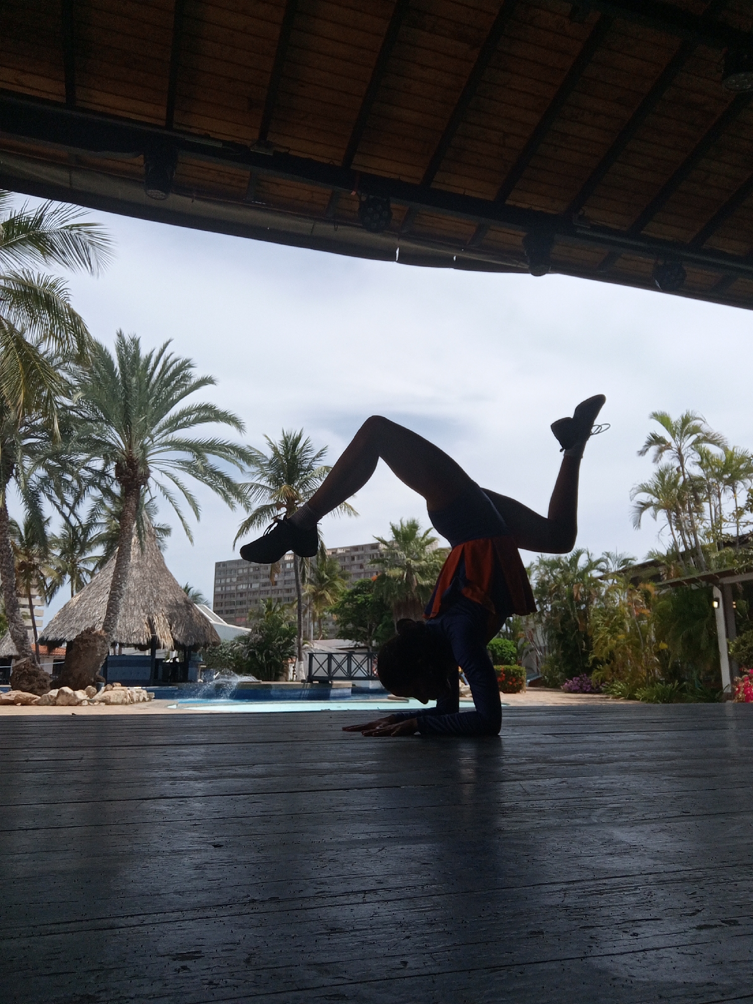 A person is performing a forearm stand on a wooden surface. The person is wearing dark athletic shoes, dark leggings, a dark long-sleeved shirt, and an orange skirt. The person's head is down towards the wooden surface.
Behind the person, there is a swimming pool, a tiki hut, palm trees, and a building. The sky is cloudy. Above the person, there is a dark wooden roof structure with lights hanging from it.
This scene appears to be in a resort area, likely in Porlamar, Venezuela, given the tropical environment and the resort-like features.