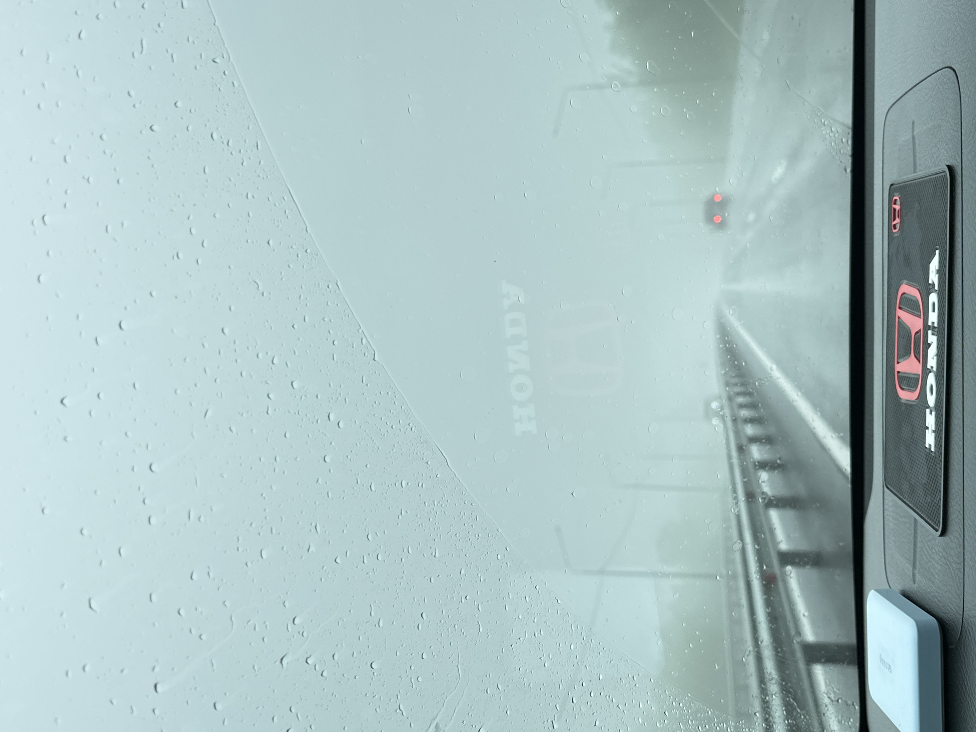 The image shows a view from inside a vehicle looking out through a rain-streaked windshield onto a highway. The weather is overcast and foggy, with visible raindrops on the glass. The road ahead is wet and appears to be a multi-lane highway with a guardrail to the left. In the distance, another vehicle with red taillights is visible. On the dashboard of the car, there is a light blue object on the left and a black mat with the Honda logo in red on the right. The Honda logo is also faintly visible on the windshield, appearing as a watermark. The overall scene suggests travel in rainy conditions on a highway. The location in the city of Пионерский, Russia, is indicated by the presence of the Honda brand, which is common in Russia, and the highway infrastructure.