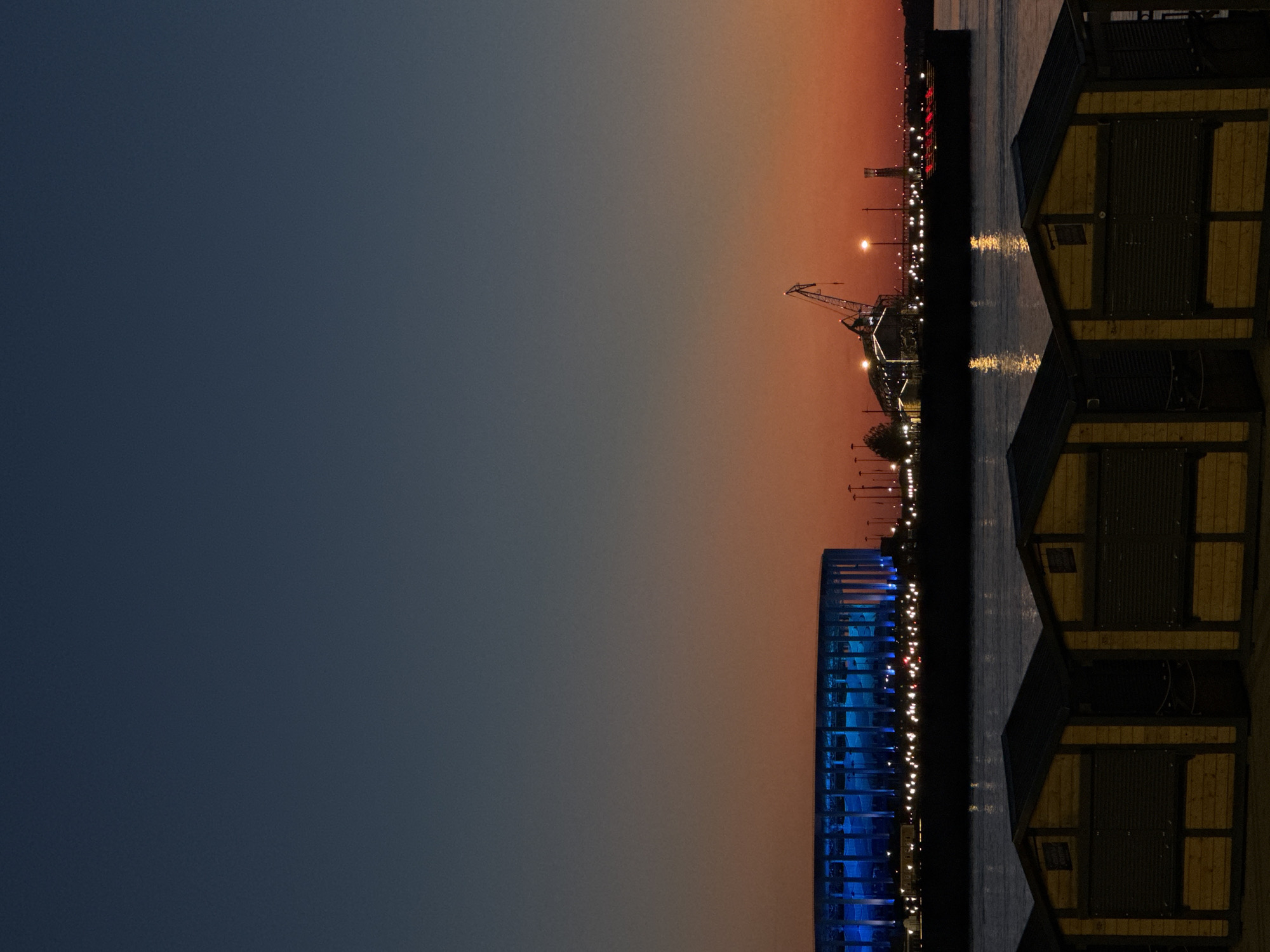 The image captures a twilight scene over a body of water, featuring a gradient sky and urban elements. The sky transitions from a dark, muted blue-grey at the top to a vibrant orange and red hue near the horizon.
In the midground, a large, circular stadium-like structure is prominent on the left, its exterior illuminated with bright blue lights. To its right, an industrial or urban waterfront area extends across the frame, characterized by numerous small, bright artificial lights that reflect on the surface of the calm water. A tall, dark crane is silhouetted against the colorful sky on the right side of this midground section.
The foreground features three horizontally aligned, small, dark wooden structures. These appear to be kiosks or stalls, each with a dark, sloping roof and closed, dark shutters covering their selling windows. The wooden paneling of these structures is faintly visible in the low light. No people or discernible actions are present in the scene. The location is an unnamed city, an unknown specific location.