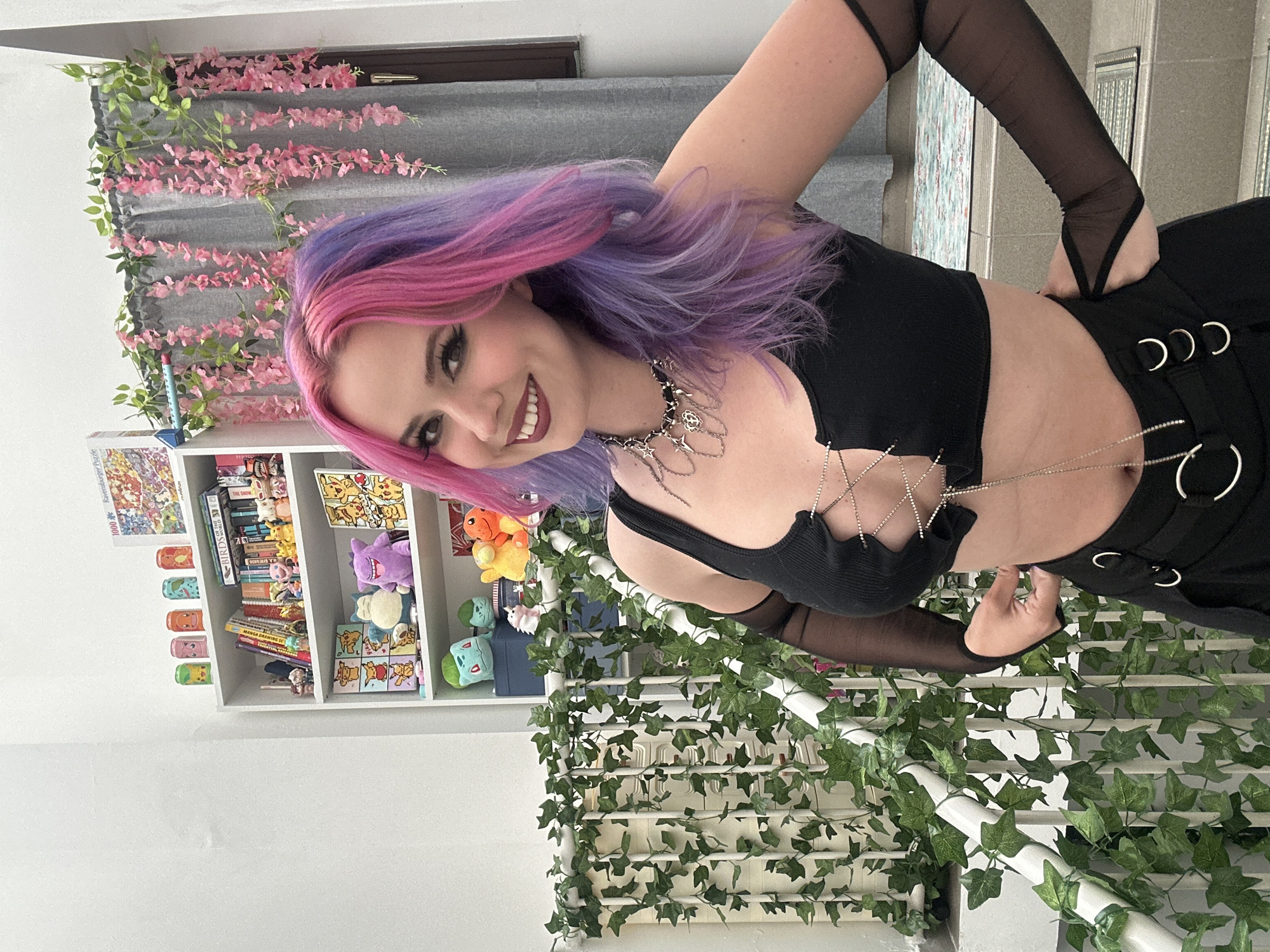 A woman with dyed purple and pink hair stands indoors next to a white bookshelf filled with books, games, and stuffed animals, as well as a white railing covered in artificial ivy. She wears a black crop top with mesh sleeves and black pants with metal rings. She is smiling and has her hand on her hip. Artificial pink flowers are draped above the bookcase. The location appears to be indoors. It is not possible to determine the specific location within the city of Zagreb, Croatia, from the image.