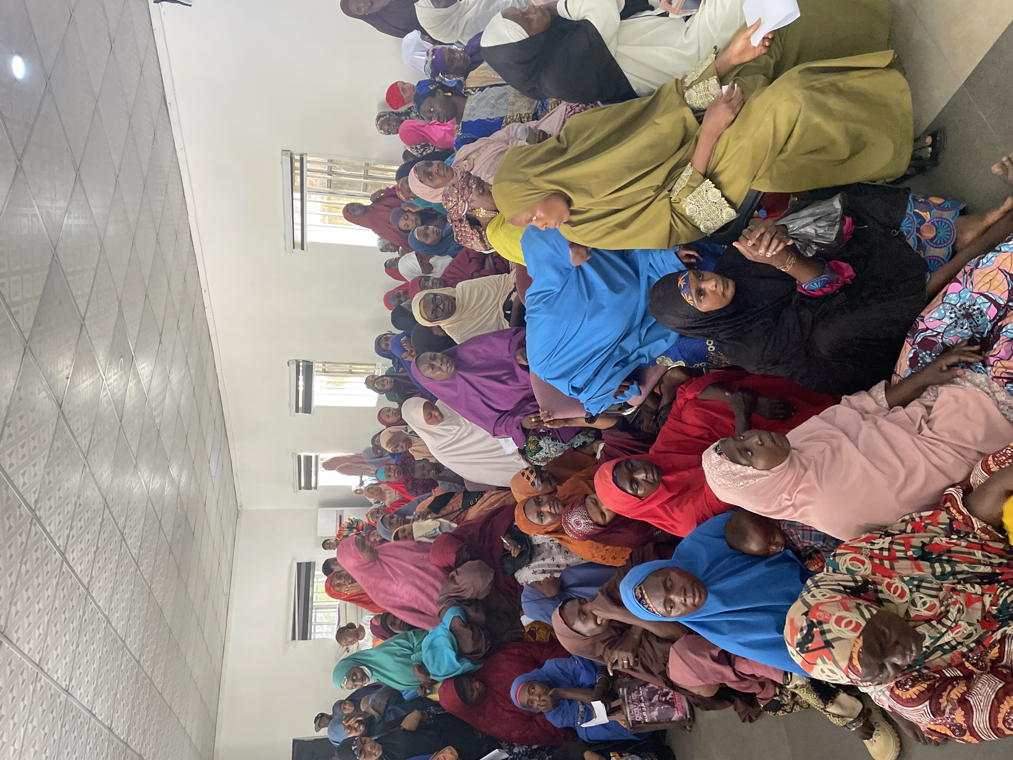 A large group of women are seated on the floor in an indoor space in Jos, Nigeria. The women are wearing clothing and head coverings of various colors, including shades of blue, red, pink, green, purple, and white. Some women are holding objects, such as papers, in their hands. The room has tile floors and windows along one side.