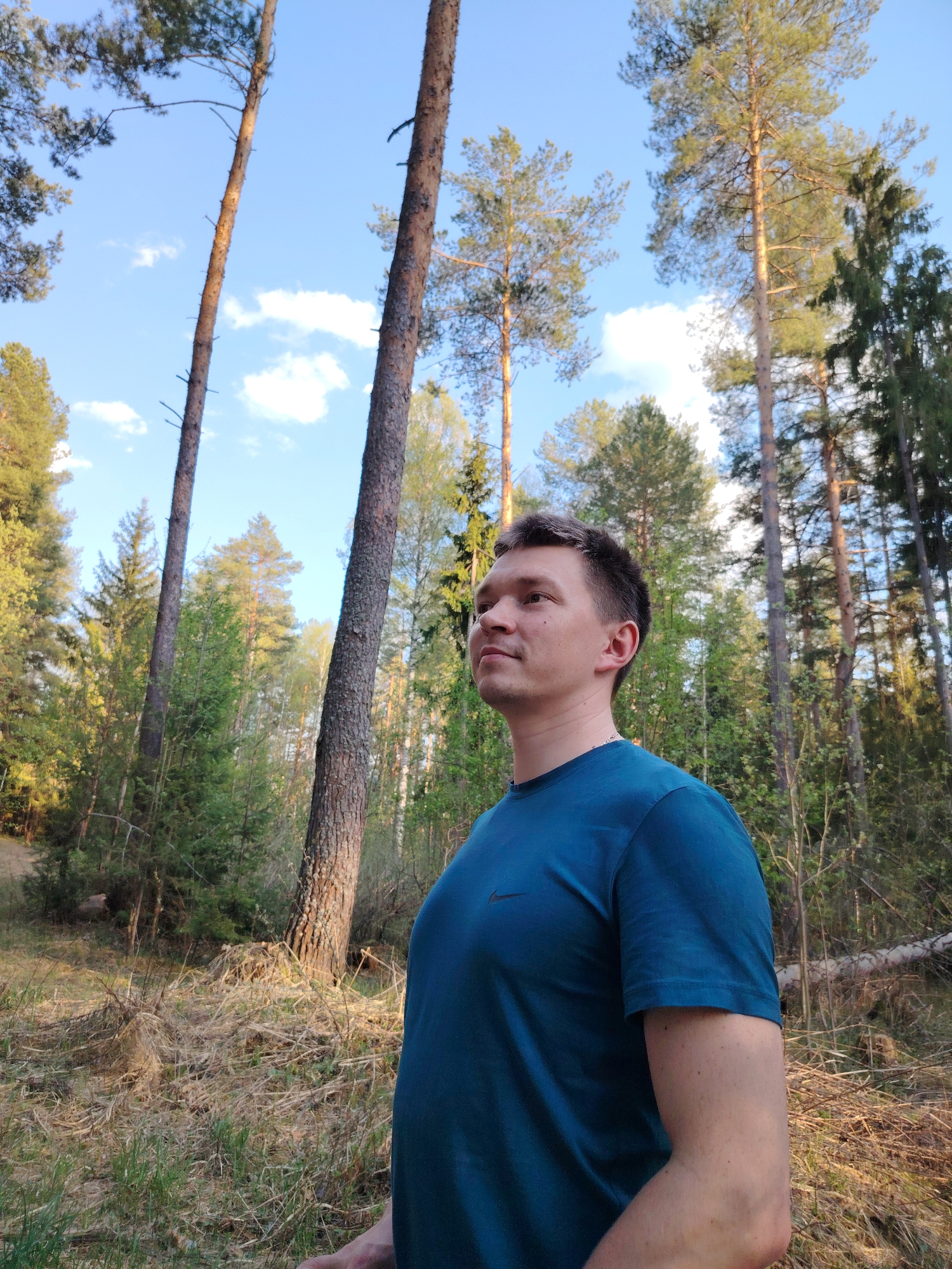 The image features a man in a blue t-shirt standing in a forest. The location is likely near Марковский, Russia, based on the prevalence of coniferous trees in the background. The man is looking slightly upwards and to the left. The background includes tall trees with a mix of coniferous and deciduous foliage. The sky is visible through the trees and has a light blue color with scattered clouds. The ground is covered in dry grass and fallen leaves. The overall lighting suggests it is daytime.