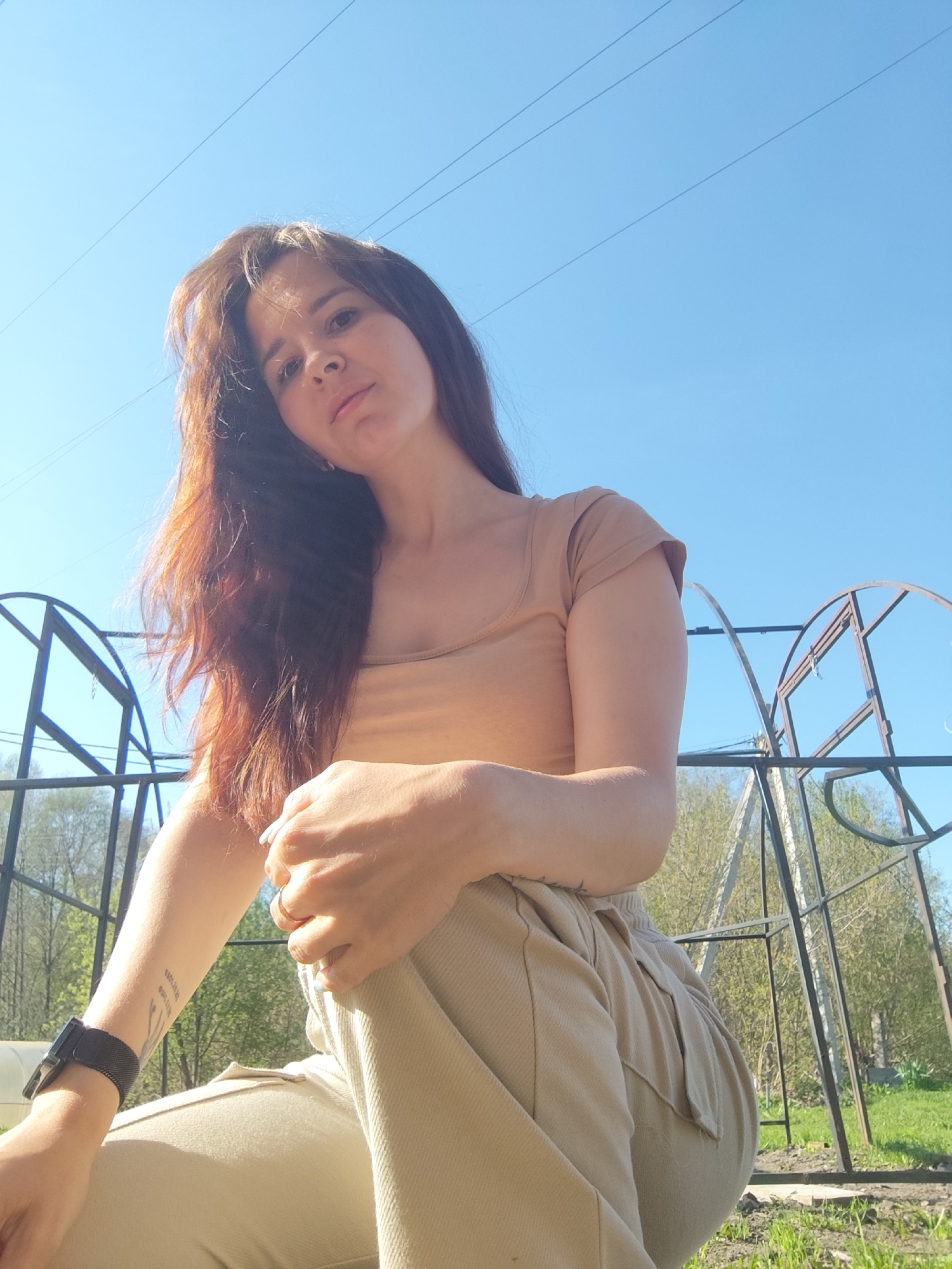 A young woman sits outdoors with a low-angle perspective under a bright blue sky. She is wearing a tan-colored t-shirt and matching pants. On her left wrist, she wears a black smartwatch and has a tattoo on her forearm. She has long, brown hair and fair skin. Behind her are metal frameworks, possibly part of a garden structure, suggesting an outdoor setting in a yard or garden. The location is in Фоки, Russia.