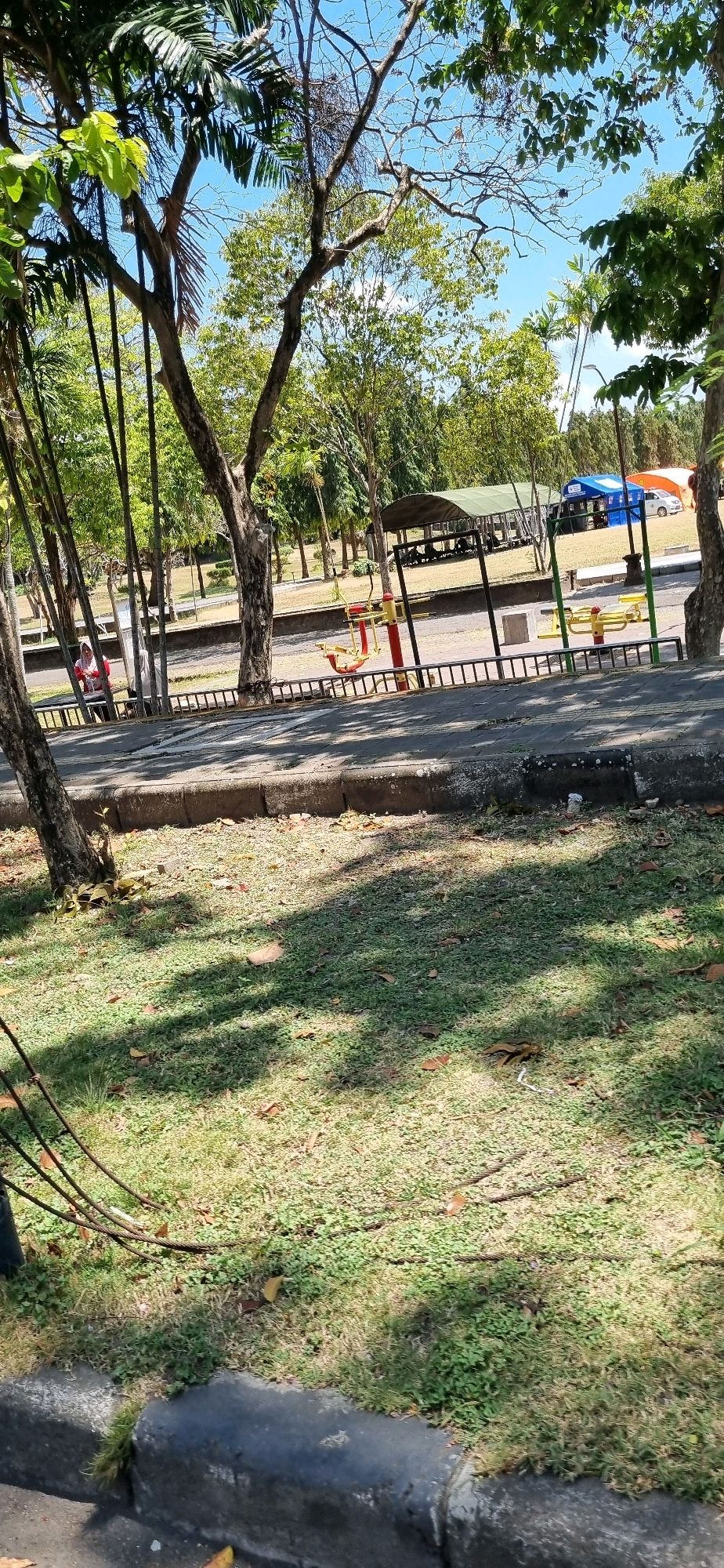 This is a photograph taken in a park or recreational area, likely in Denpasar, Indonesia, given the tropical vegetation and climate. In the foreground, a paved walkway runs along a grassy area with scattered dry leaves. Beyond the walkway, there are several pieces of outdoor exercise equipment, including yellow and red machines. A black metal fence separates the walkway from the area with the exercise equipment and a paved road.
In the middle ground, there are several large trees with lush green foliage. A covered pavilion or shelter with a green roof is visible, and under it, several people are seated, possibly resting or picnicking. Further in the background, a white car is parked near two tents, one blue and one orange, suggesting a temporary event or gathering. The sky is clear and blue, indicating fair weather. The overall impression is of a public space designed for recreation and relaxation, with elements of nature and man-made structures coexisting.