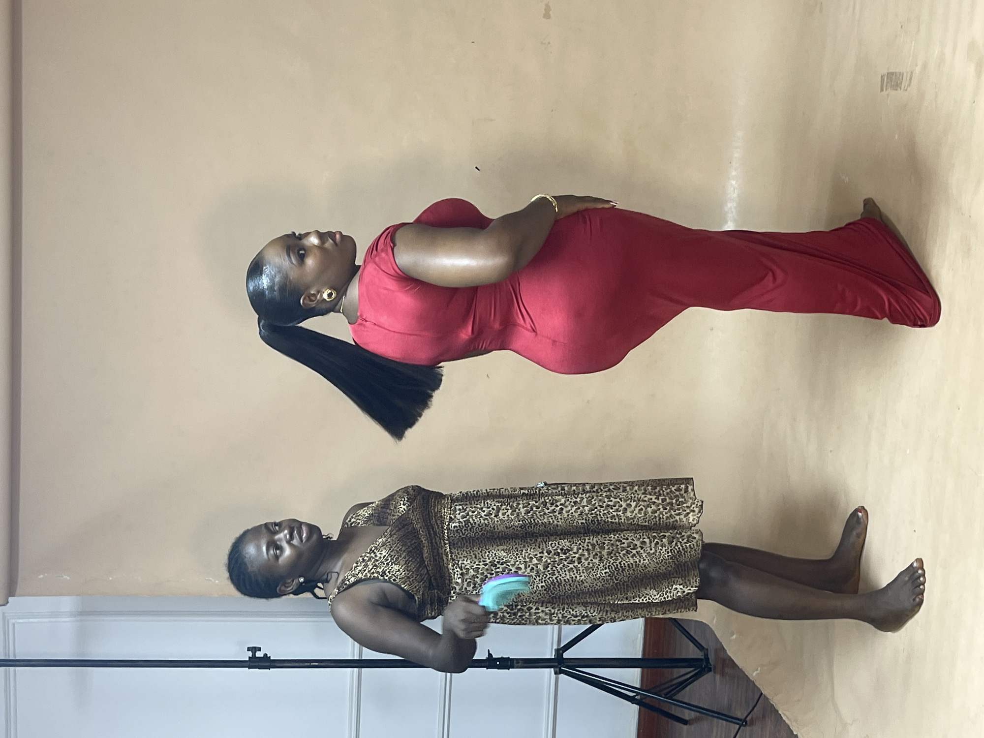 The image depicts two women in what appears to be a studio setting. The backdrop is a seamless beige paper. One woman is lying in a red dress with a ponytail. The other woman is lying in a leopard print dress, holding a small object. A stand is visible. Location cannot be confirmed as Abuja, Nigeria.