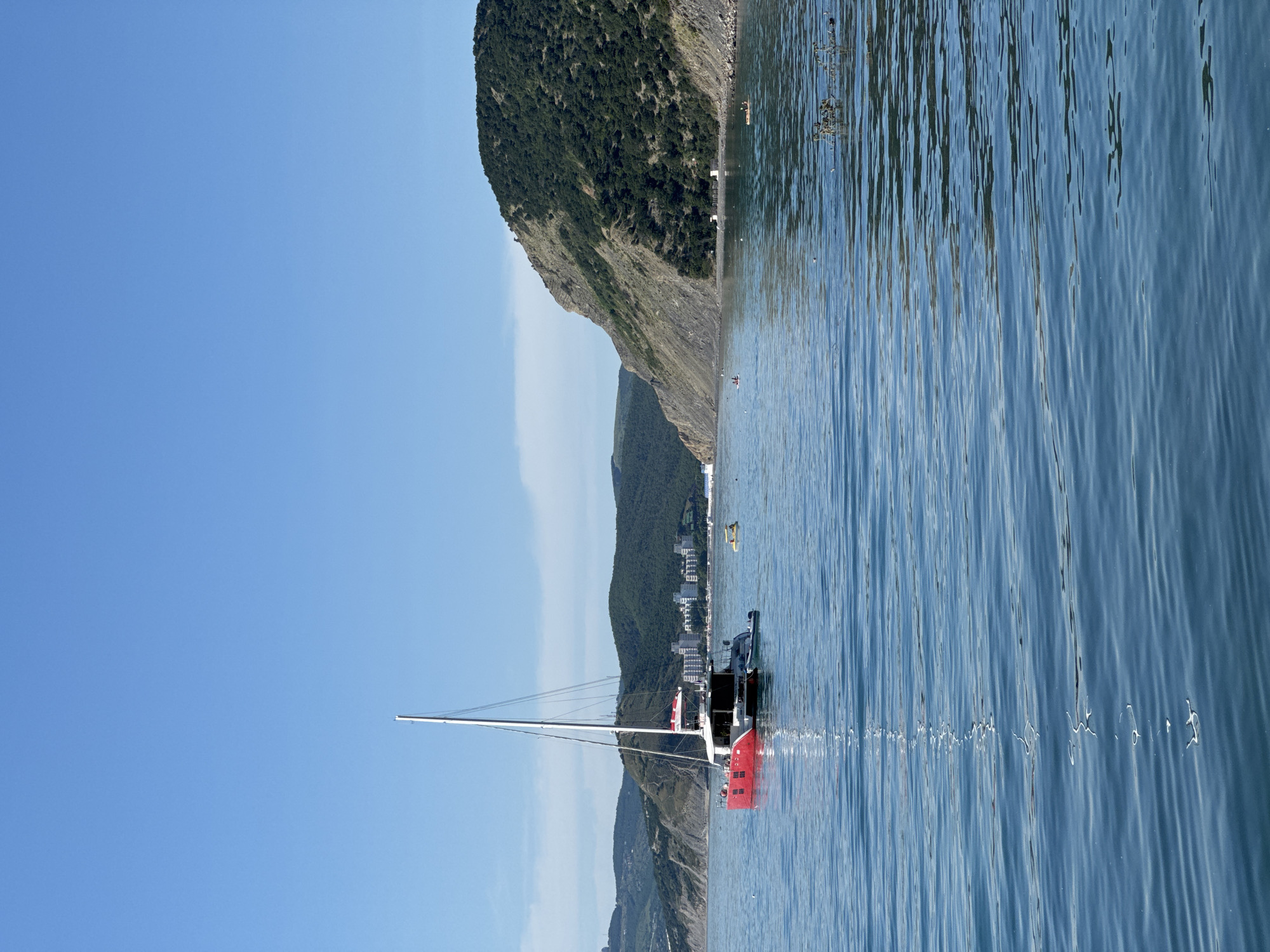 This serene coastal scene, captured in Bol'shoi Utrish, Russia, depicts a bright, clear day over calm blue waters. The primary subject is a large sailboat, possibly a catamaran, positioned slightly to the left of the frame. It features a tall white mast, a predominantly white hull, and a distinctive red section on its side. A flag, appearing red and white, is visible at its stern.
In the background, a dramatic coastline rises with steep, green-covered hills and mountains descending directly into the sea. Along the base of these hills, a scattering of white and light-colored buildings suggests a small town or resort nestled by the shore. Several small vessels, including a yellow kayak and another white one, are dotted across the water closer to the coast, indicating leisurely activity. The water itself is calm with gentle ripples, reflecting the clear blue sky above, which is entirely free of clouds. The overall atmosphere is peaceful and tranquil, characteristic of a pleasant day for sailing or coastal recreation. No discernible text is visible within the image.