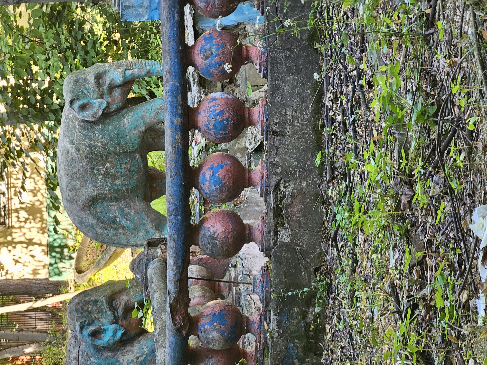 This is a medium-shot photograph of dilapidated concrete elephant statues in an outdoor setting. In the foreground, a weathered blue and red balustrade with spherical pillars runs horizontally across the frame. The balustrade is cracked and peeling. Behind the balustrade, two concrete elephant statues are visible. The left statue is partially obscured and appears to be facing left, with teal paint on its head. The right statue is a full view of an elephant standing, facing right. Both elephants are covered in chipped teal and grey paint, with some pink showing through on the right statue's trunk and ears. The ground in front of the balustrade is covered with dry leaves, twigs, and small green plants. In the background, lush green foliage and trees are visible, along with parts of a light-colored building. The overall impression is one of neglect and abandonment. This scene is located in the abandoned section of the Likhachev Park in Khimki, Russia, specifically in the area where the former "Park of Giants" was situated.