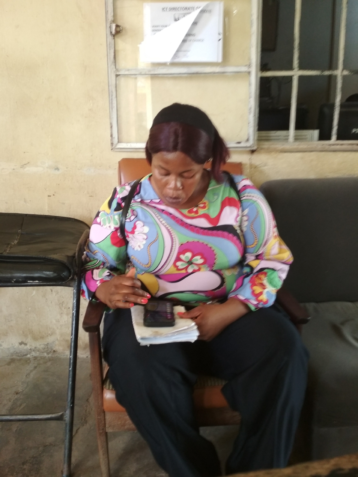 A medium shot captures a Black woman with dark hair pulled back, wearing a colorful, patterned long-sleeved blouse and black pants, seated in a wooden chair. She is looking down at a phone held in her hands, which rests on a stack of papers. The background shows a wall with a window and a bulletin board. The scene is likely indoors, possibly an office or waiting area, in Bisichi, Nigeria.
