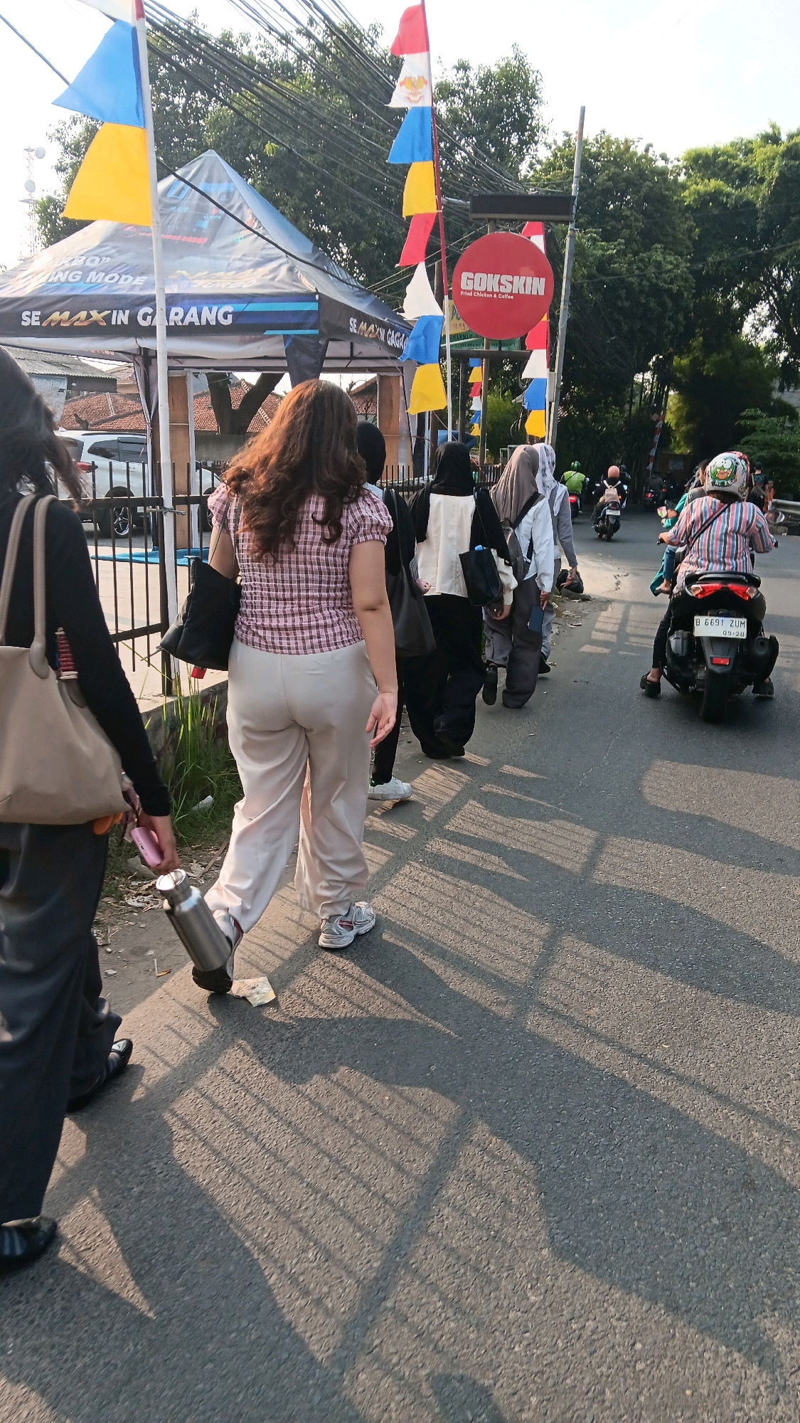 The image shows a street scene in South Jakarta, Indonesia, likely during the day with sunlight casting long shadows. In the foreground, a woman with long, brown hair is walking away from the camera, wearing a checkered pink and white short-sleeved shirt and light-colored pants. She is carrying a black bag over her left shoulder and a stainless steel water bottle in her right hand. To her left, another person, mostly out of frame, is wearing a dark long-sleeved shirt and dark pants, and carrying a large beige tote bag.
Behind the first woman, a line of people is walking along the sidewalk. Some are wearing hijabs. A red circular sign with "GOKSKIN Fried Chicken & Coffee" is visible in the background, along with a tent with banners that include the text "SE MAX IN GARANG". Several colorful flags (red, white, yellow, blue) are strung along a pole.
On the road, several motorbikes are present. One motorbike in the middle ground has a rider wearing a striped shirt and helmet, and a license plate that reads "B 6691 ZUM". The overall environment suggests a commercial or event area with people walking and motorbikes on the road, characteristic of a busy urban setting in Indonesia.