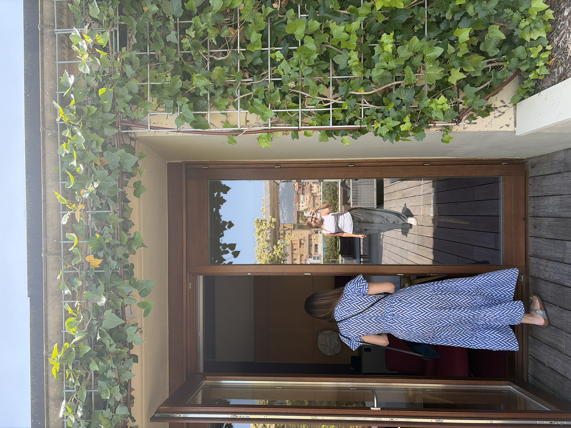 The image depicts an open glass door or window onto a wooden deck. A woman wearing a blue and white patterned dress stands partially within the interior space. Another woman can be seen reflected in the glass, holding a phone and facing outward. Vines grow on a trellis outside the window, partially obscuring the view beyond. The reflection in the glass reveals a sunny day in a city, with buildings visible in the background.