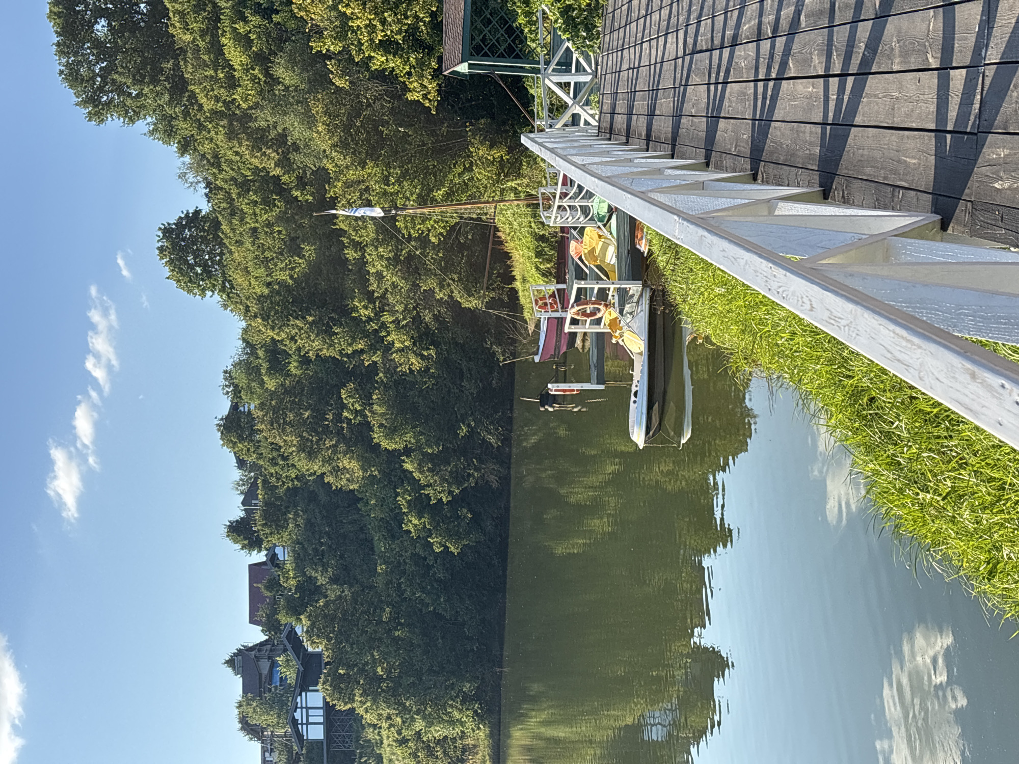 The image depicts a wooden pier extending into a calm body of water, likely a lake or river, in Romashkovo, Russia. The pier is made of dark, weathered wooden planks and has a white railing along its edge. To the left of the pier, the water reflects the surrounding lush green trees and a bright blue sky with scattered white clouds. Along the bank, vibrant green grass grows.
In the water, several small boats are docked. One boat, partially visible, is red with a white interior. Another is a white, flat-bottomed vessel. A sailboat with a tall mast and a white flag is also moored. The boats appear to be recreational.
In the background, a dense forest covers a hillside. Several houses are visible nestled amongst the trees on the upper part of the hill, suggesting a residential area. A small, green, open-sided structure with a sloped roof is located near the end of the pier. The overall scene suggests a tranquil, natural setting within or near a populated area. There are no people visible in the image.