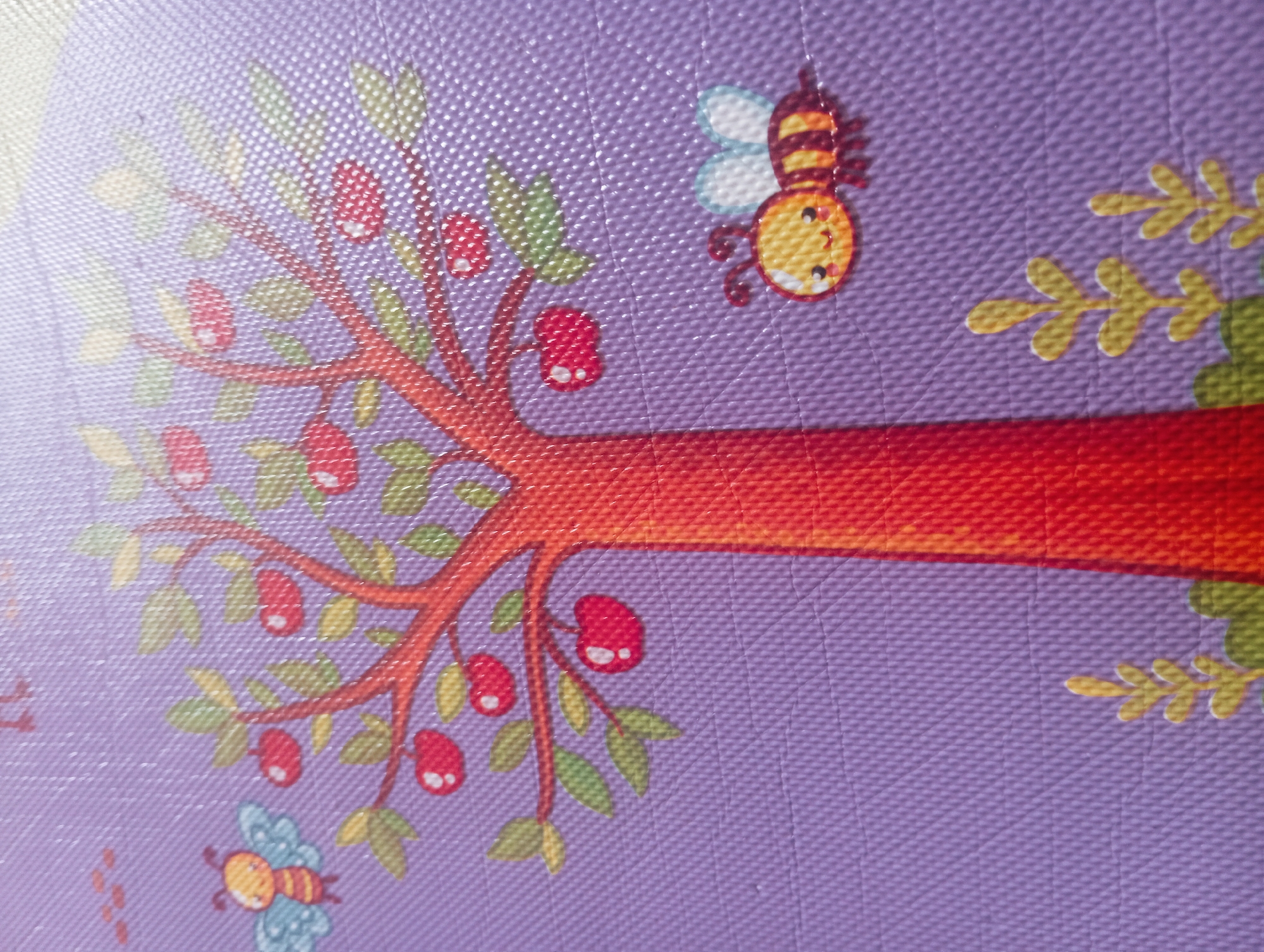This is a close-up, overhead shot of a child's play mat. The mat features a cartoon illustration of an apple tree with red apples and green leaves against a lavender background. Two cartoon bees are also depicted: one flying near the top left of the tree, and another to the right of the tree, near the bottom. The surface of the mat has a textured, bumpy pattern. There are no people or indications of a specific location like Kutasari, Indonesia.