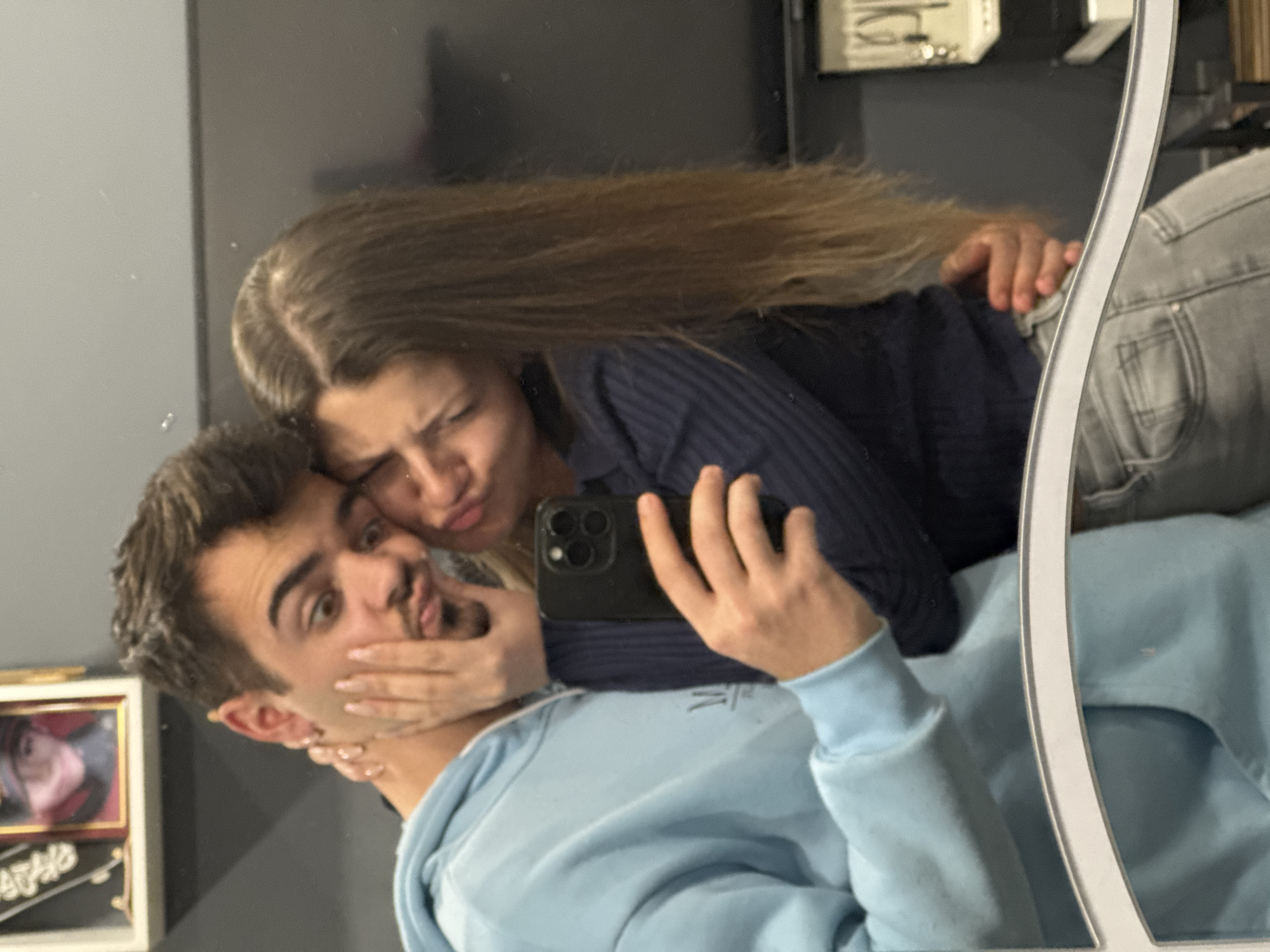 A selfie of two people in front of a mirror. A light-skinned male with short, dark hair is wearing a light blue hoodie. A light-skinned female with long, blonde hair is wearing a dark blue top and light gray jeans. The woman is holding a dark-colored phone with multiple camera lenses. The background includes a gray wall with a framed picture and other objects on shelves. The location is possibly indoors, but the city of Sofia, Bulgaria cannot be confirmed from the image.