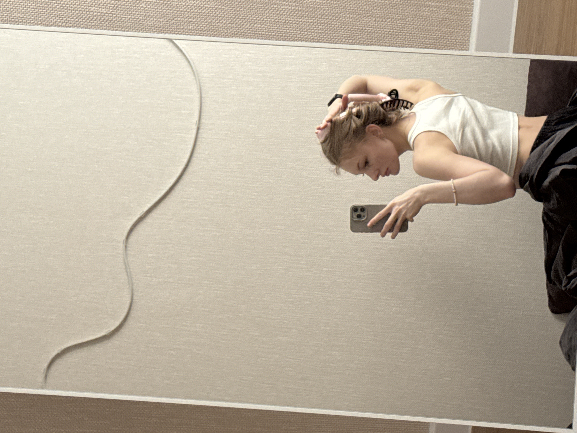A young woman is reflected in a mirror. She is holding a smartphone in her right hand, appearing to take a photograph or video of herself. She is wearing a white tank top and dark pants, and her hair is styled with rollers or curlers. The mirror hangs on a wall with a textured beige or light brown surface. A decorative line or pattern is visible on the wall behind the mirror. The location is in Perm, Russia.