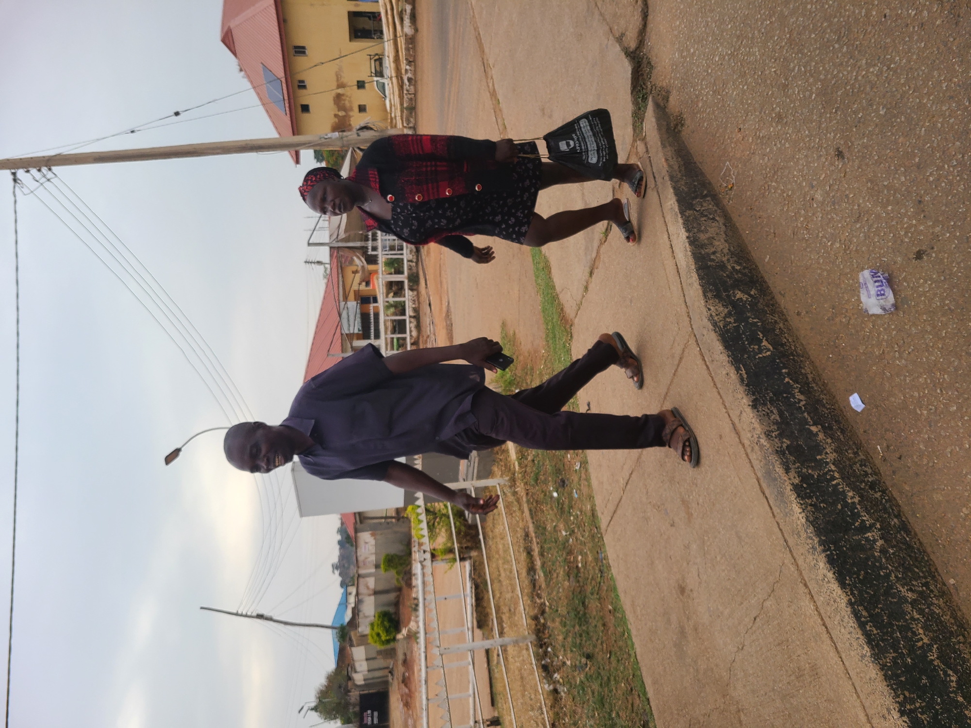 The image captures an outdoor scene in what appears to be an urban or semi-urban area of Jos, Nigeria. The photograph is oriented sideways, showing two individuals walking along a concrete sidewalk beside a road.
In the foreground, a man with short dark hair walks towards the left of the frame (when viewed upright). He is dressed in a dark blue short-sleeved shirt, dark trousers, and dark sandals, and holds a dark-colored smartphone in his left hand. His expression is neutral. Behind him and slightly to his right, a woman is also walking. She wears a black dress with a light pattern, a red and black plaid jacket or cardigan, and a patterned head covering. She carries a black tote bag over her left shoulder and wears sandals. Her expression is also neutral.
The setting includes a paved sidewalk with a visible curb separating it from the adjacent ground. The ground cover consists of small stones and dirt, with some sparse patches of grass and weeds. In the background, several buildings are visible; one prominent structure has a yellow facade and a red roof, featuring what looks like a solar panel. Other buildings with red and blue roofs can be seen further back. A wooden utility pole with multiple overhead wires stretches across the upper left portion of the image, and a street light fixture is also visible in the background. The sky is bright but appears overcast or hazy, suggesting it is daytime, possibly late morning or afternoon.
Notable details include a crumpled white piece of packaging on the ground to the right, near the curb, with the blue letters "BUM" clearly visible. The black tote bag carried by the woman has some white text and what might be a vehicle icon, but the full text is indistinct. The overall impression is of a regular day in a bustling street environment.