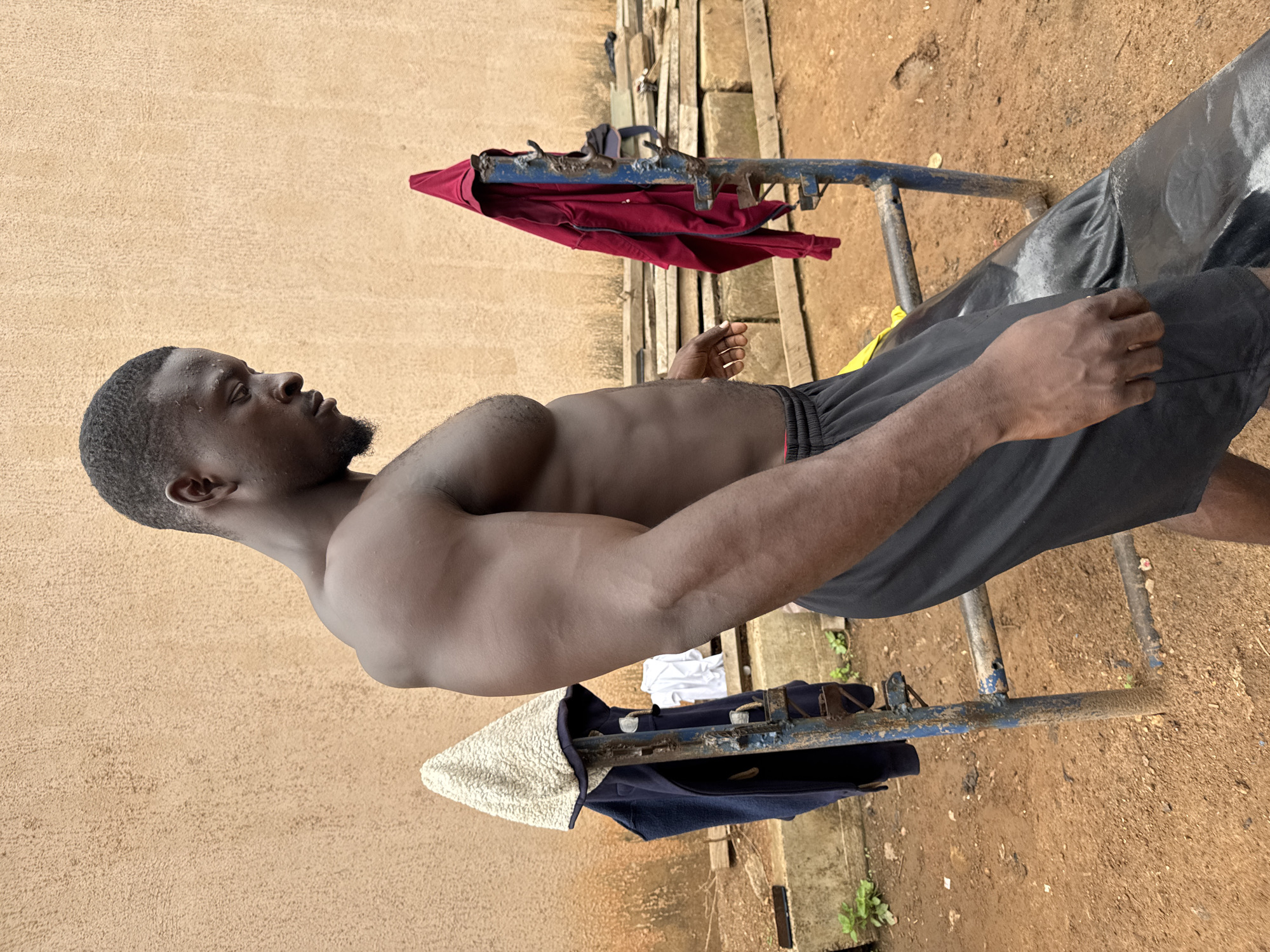 A medium shot captures a man in an outdoor setting, seemingly in Akwanga, Nigeria, given the background environment. He is partially undressed, wearing black shorts. The man is positioned in front of a blue, weathered metal structure, which appears to be a makeshift clothes rack. Several articles of clothing, including a red shirt, are draped over the rack. The ground is dirt with some vegetation. A wall is visible on the left side of the frame.