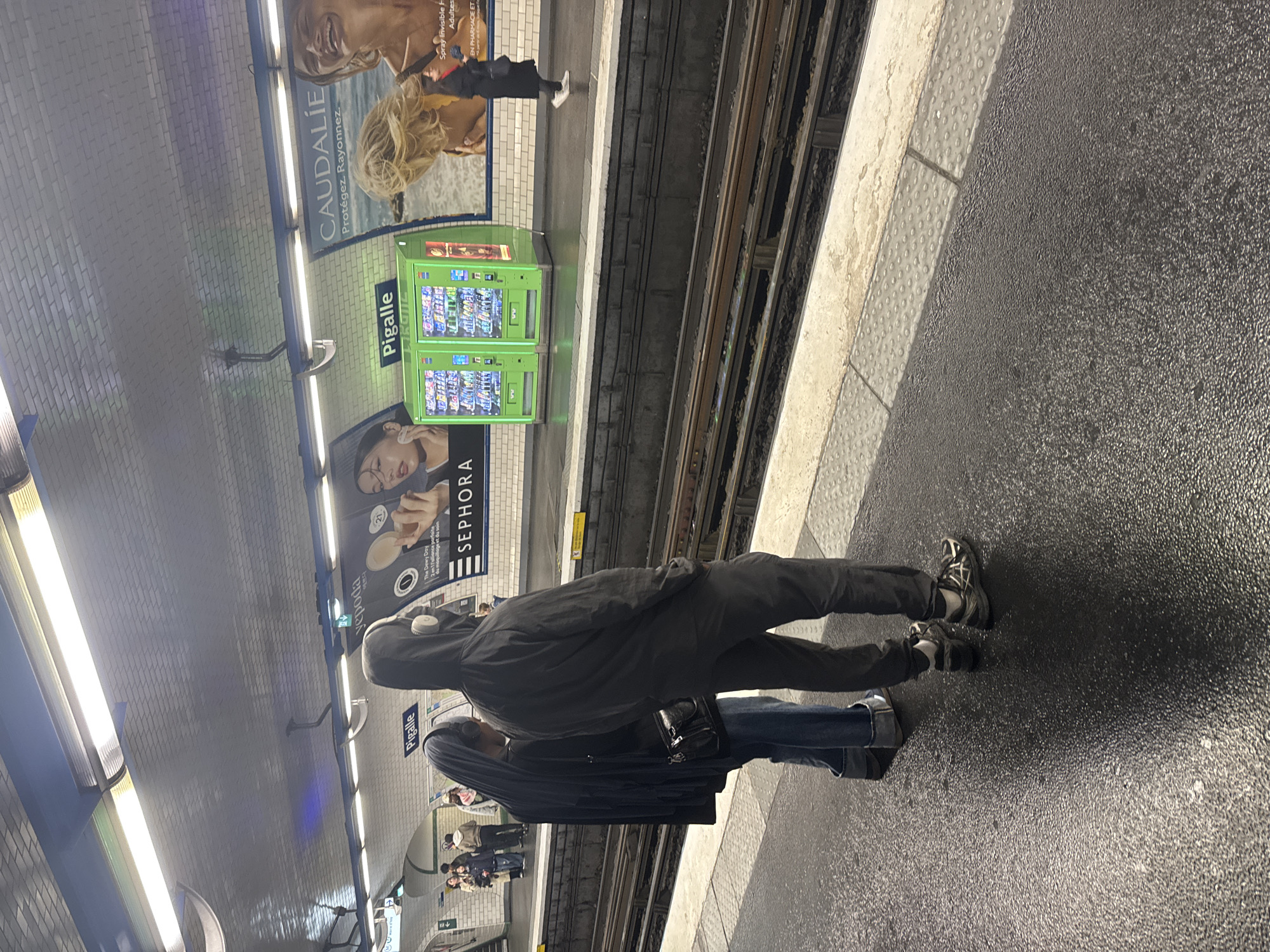 Here is a description of the image:
This is a medium shot of a subway platform at the Pigalle station in Paris, France. Two individuals stand near the edge of the platform, adjacent to the subway tracks. The person closer to the tracks is wearing a dark hooded sweatshirt and trousers with athletic shoes. This person has headphones on their head. Beside them is someone wearing jeans and a black coat, carrying a small black handbag. Subway tiles, advertisements for Caudalie and Sephora, and vending machines can be seen along the walls. Fluorescent lights illuminate the scene. Other people are present further down the platform. The platform surface is dark and textured.