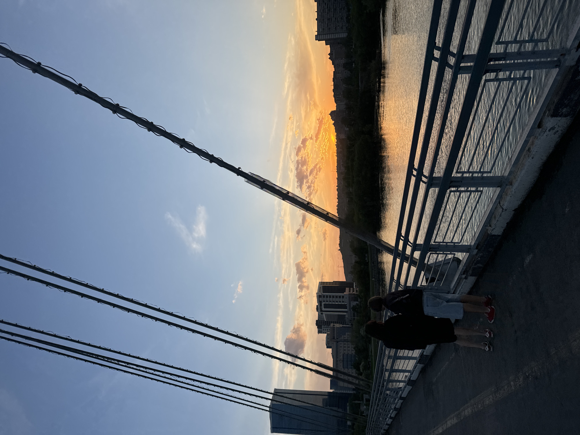 Two people stand on a bridge next to a metal railing. In the background is a river and buildings silhouetted against a colorful sunset. Power lines are visible in the foreground. This is the Vinogradovsky Bridge in Krasnoyarsk, Russia.