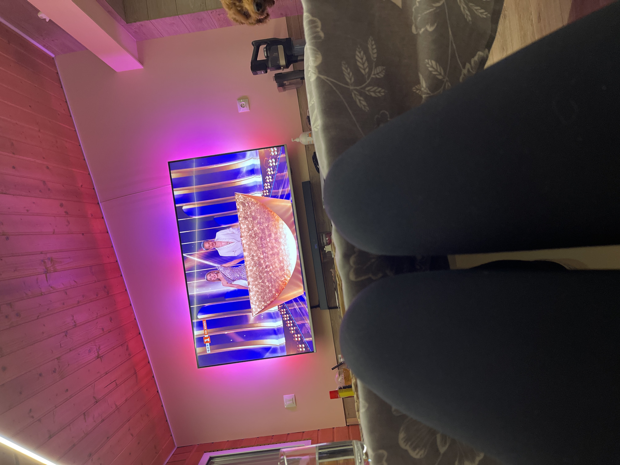 The image shows a living room interior with a television displaying a show. The room has a slanted wooden ceiling illuminated with pink light. A couch with a floral patterned cover is present. In the foreground, a person's legs, clad in dark leggings, obscure the view. A vacuum cleaner is mounted on the wall. Bottles and glasses are on a table. Although the television broadcast is in Russian, there is no information to determine if this is in the city of Sofia, Bulgaria.