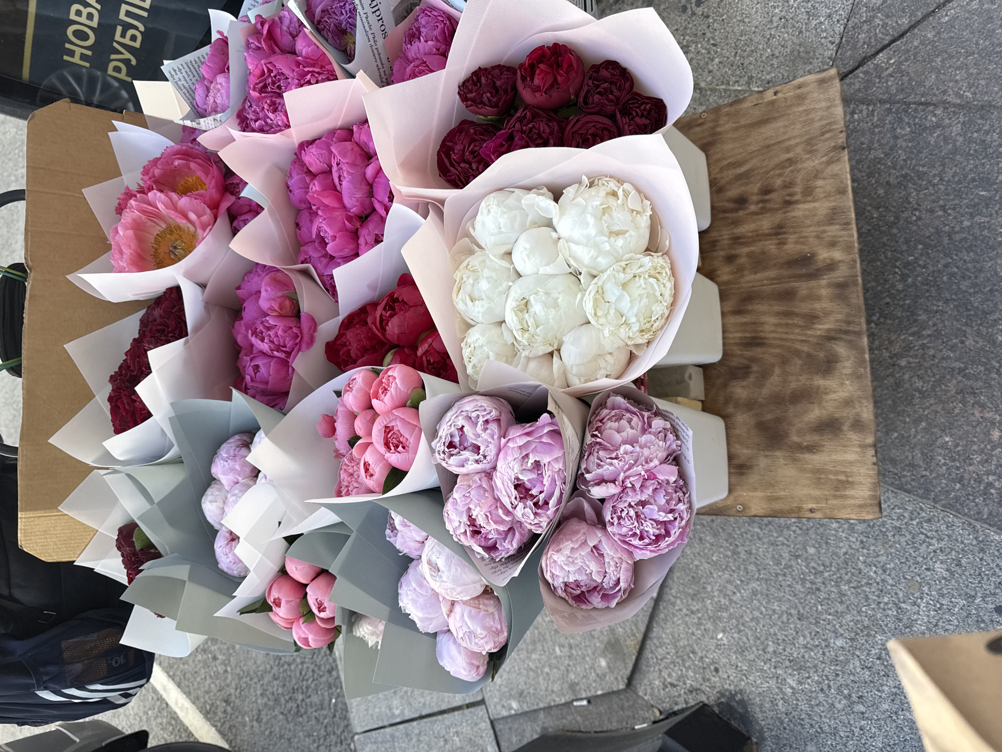 The image shows a close-up, eye-level view of several bouquets of peonies arranged on a wooden stand on a city sidewalk. The peonies are in various stages of bloom and come in colors including light pink, deep pink, white, and dark red. Each bouquet is wrapped in paper, some of which is white, light pink, or light blue-grey. In the background, a brown cardboard box is visible, along with the edge of a dark backpack or bag with white stripes. The sidewalk is paved with grey cobblestones. A portion of a sign with Cyrillic text is visible in the upper right corner, reading "НОВЫ" and "РУБЛЬ", indicating a location in Saint Petersburg, Russia, likely a flower stand or market. No people are visible in the frame, and the flowers appear to be stationary. The lighting suggests it is daytime.
