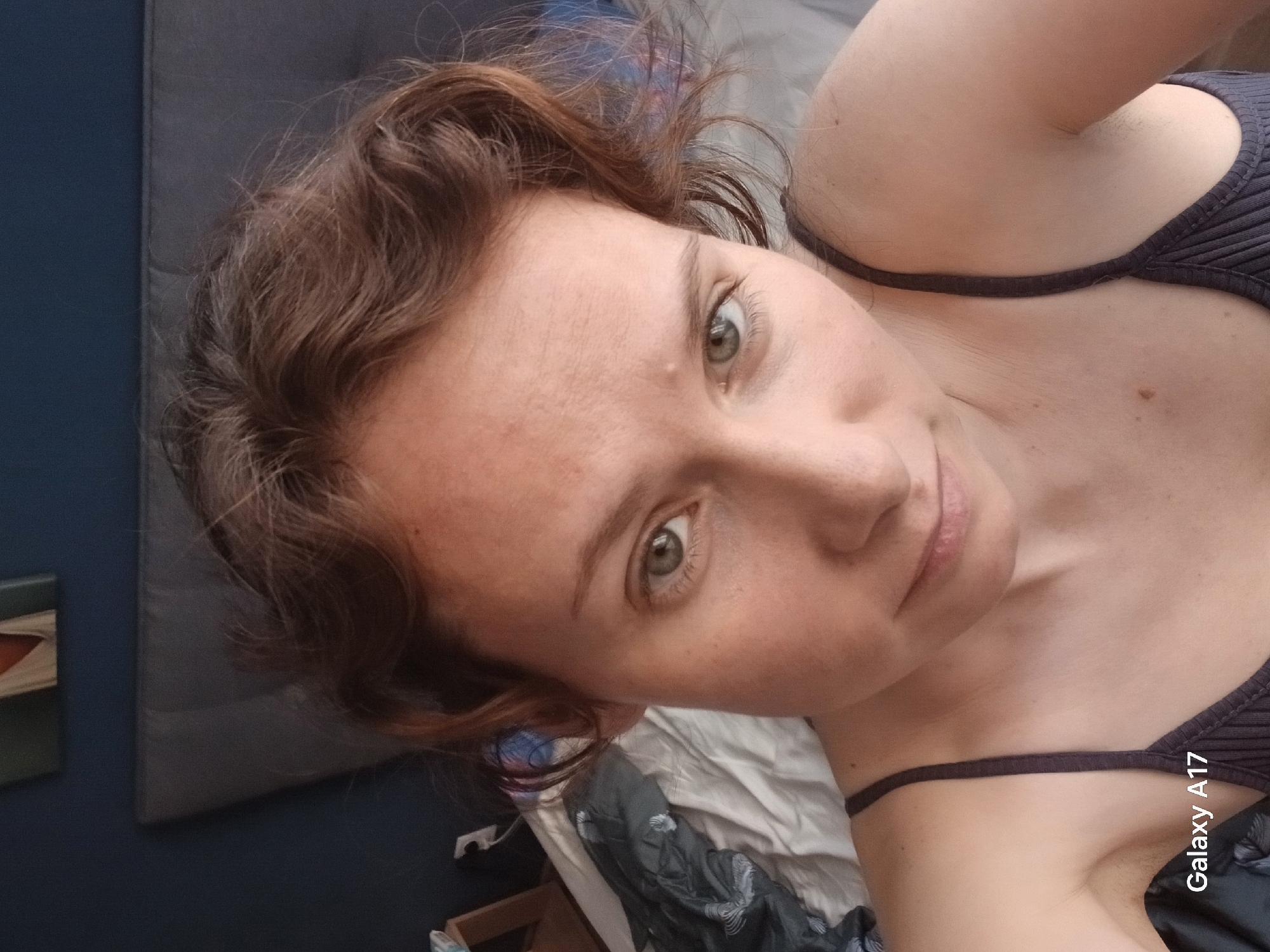 A woman with reddish-brown wavy hair is facing the camera with a slightly upward gaze. She has fair skin with some freckles. She's wearing a dark-colored, possibly ribbed, tank top or sports bra with thin straps. The background includes a blue wall, a gray cushioned headboard, and crumpled white and dark-colored bedding. A "Galaxy A17" watermark is visible in the lower right corner. The location, Yekaterinburg, Russia, cannot be determined from this image alone.