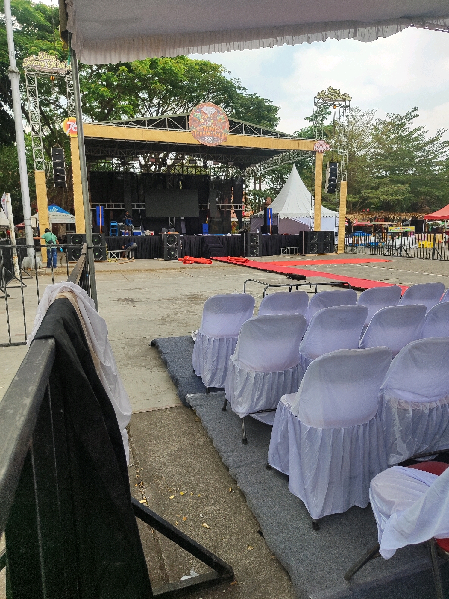 The image depicts an outdoor event setup, likely in the city of Trenggalek, Indonesia, given the signage. In the foreground, rows of chairs are covered with white fabric, some resting on grey carpeted mats. Behind these chairs is a concrete area with a black metal fence and some discarded fabric. Further back, a stage is set up with a large LED screen and audio equipment. A red carpet runner extends from the stage area. Various tents, including a white peaked tent, are visible behind the stage, along with trees and a cloudy sky. Two individuals are seen working on the stage. One is standing with a bag, and another is bent over near some wooden planks. The scene suggests preparations for a public gathering or performance.