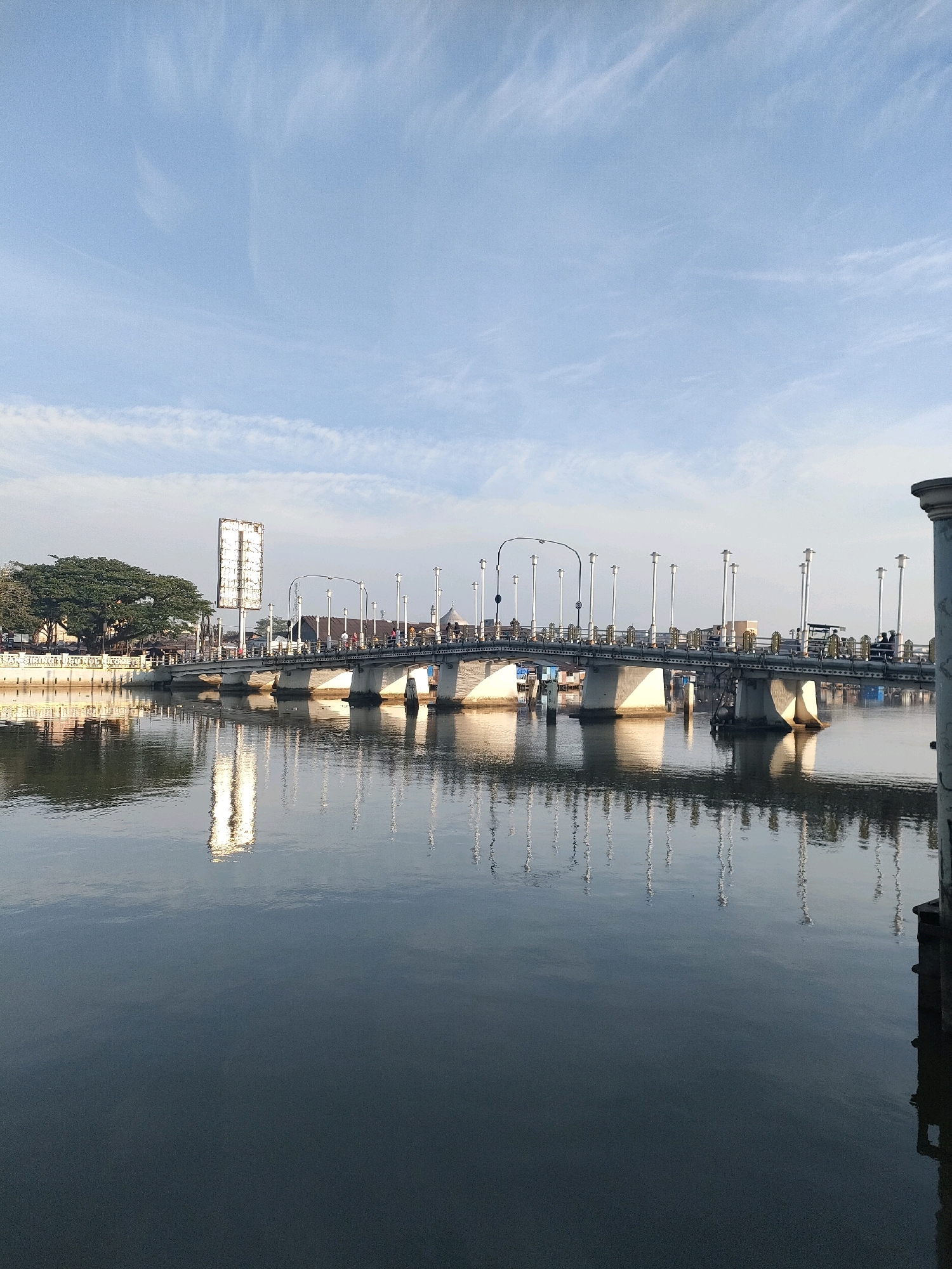 The image depicts a bridge spanning a body of water in Banjarmasin, Indonesia. The bridge, a concrete structure with lampposts lining its railings, reflects in the calm water below. On the left bank, a large tree and a billboard are visible. The sky is a clear blue with wispy clouds. The overall scene suggests a serene urban landscape during daylight hours.
