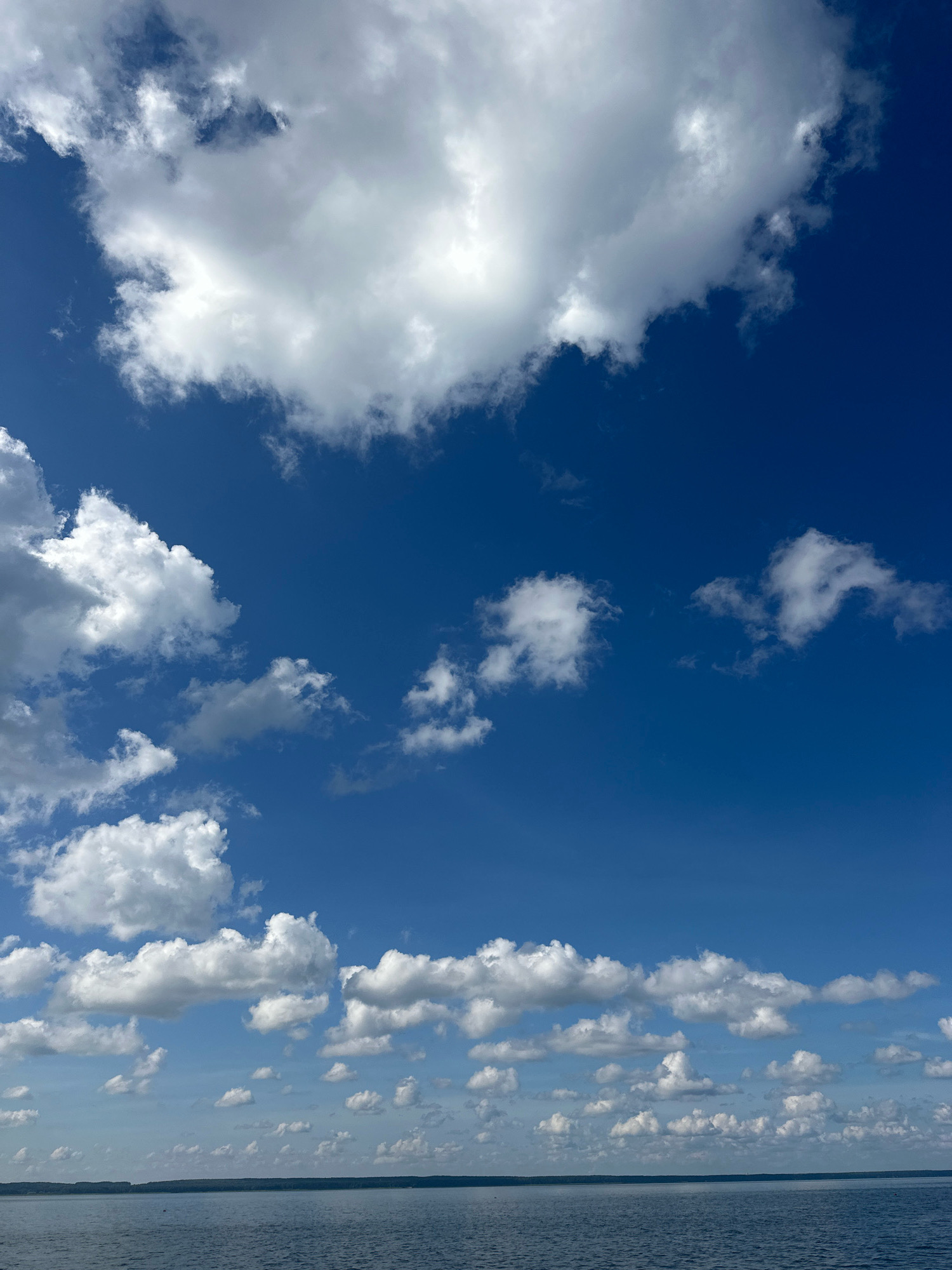 The image depicts a vast expanse of deep blue sky dotted with numerous white, fluffy cumulus clouds. The clouds vary in size and density, with some appearing as large, dramatic formations and others as smaller, scattered wisps. The lower portion of the image shows a calm body of water, likely a lake or river, with a distant, dark green shoreline visible on the horizon. The overall impression is one of a clear, bright day with scattered clouds. The location is identified as Мядзельскі сельскі Савет, Belarus. There are no people, animals, or man-made objects visible in the image. The scene is purely natural, focusing on the sky and water.
