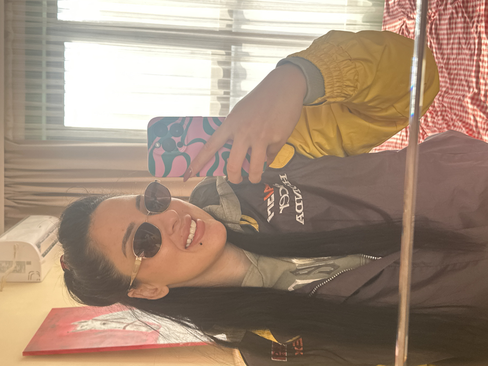 A person is taking a selfie in a mirror. The person is wearing sunglasses and a brown jacket with yellow sleeves. They are holding a phone with a pink and green patterned case. The background includes a window with blinds, a red and white checkered curtain, a painting, and a white object. The location cannot be determined as being in Varna, Bulgaria from this image.