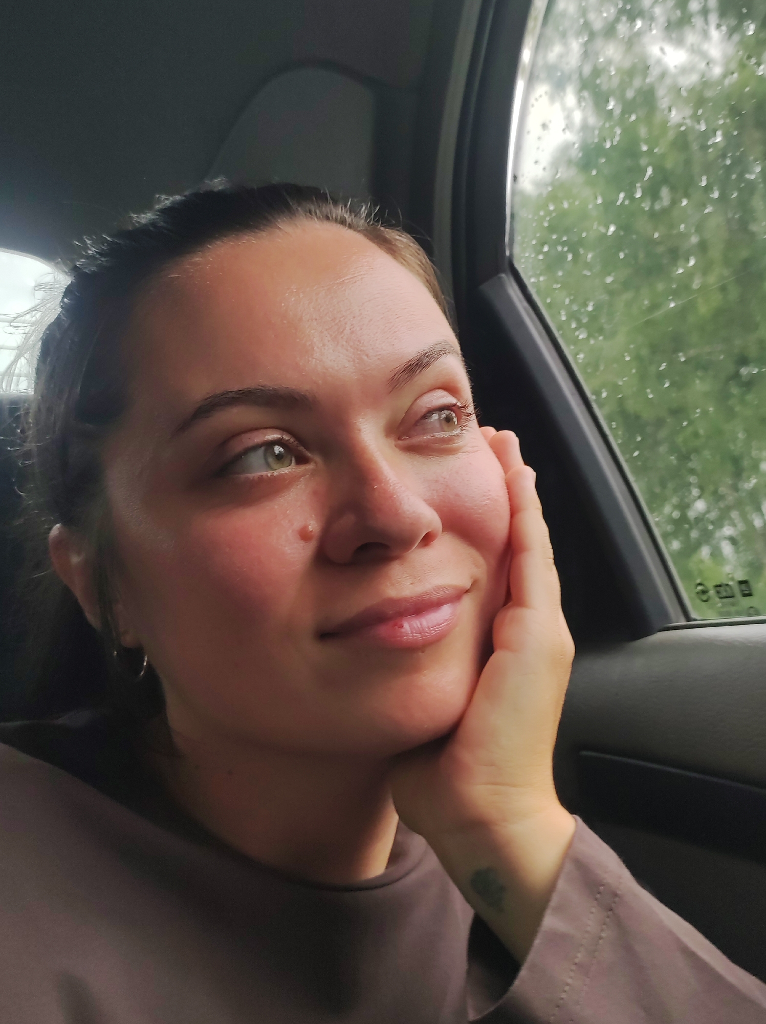 A medium shot captures a young woman in the passenger seat of a car, looking out the window at a rainy, green, outdoor scene. Her right hand rests against her cheek, and her gaze is directed upwards and to the right. Her hair is pulled back, and she is wearing a dark brown top. Raindrops are visible on the car window, blurring the background. The interior of the car, including the door panel and window frame, is visible in the foreground. The woman has a mole on her right cheek and a small tattoo on her left wrist. The image was taken in Chaikovsky, Russia.