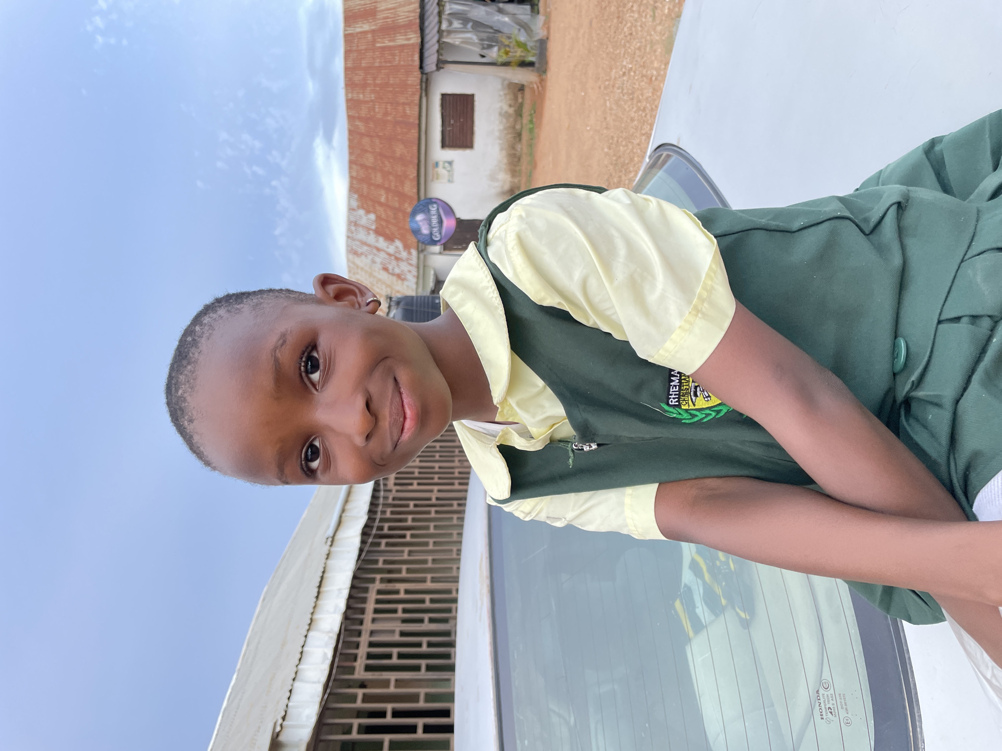 A young girl in a school uniform with the inscription "RHEMA CHRISTIAN" is sitting on the hood of a silver-colored car. She has short, dark hair and is smiling. Behind her are buildings with corrugated iron roofs. A sign for "GOLDBERG" is visible in the background. The location is Barakin Akawo, Nigeria. The sky is overcast. A security door is visible to the left side.