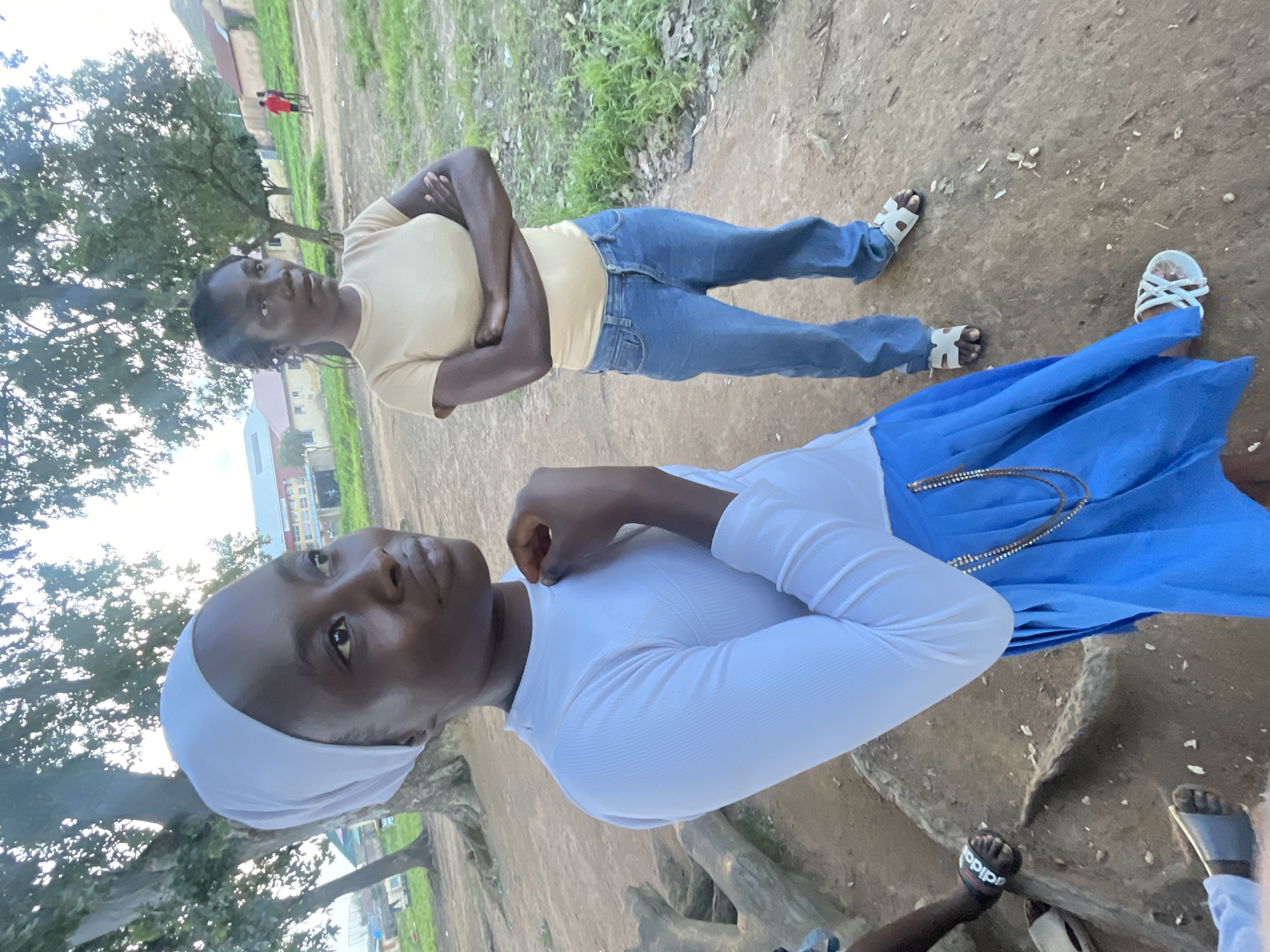 In Jos, Nigeria, two young women are captured in a medium shot outdoors, with a focus on the woman in the foreground. She is wearing a white long-sleeved shirt and a bright blue skirt, adorned with a beaded necklace. Her head is covered with a white headscarf. She is looking to her right, with her hand raised to her neck. Behind her, a second woman stands with her arms crossed, wearing a light yellow t-shirt and blue jeans, looking off to her left. The background reveals a rustic environment with dirt ground, scattered debris, and sparse vegetation. Trees are visible, and in the distance, buildings with red roofs suggest a settlement. The lighting indicates it is daytime, possibly late afternoon, with soft natural light.