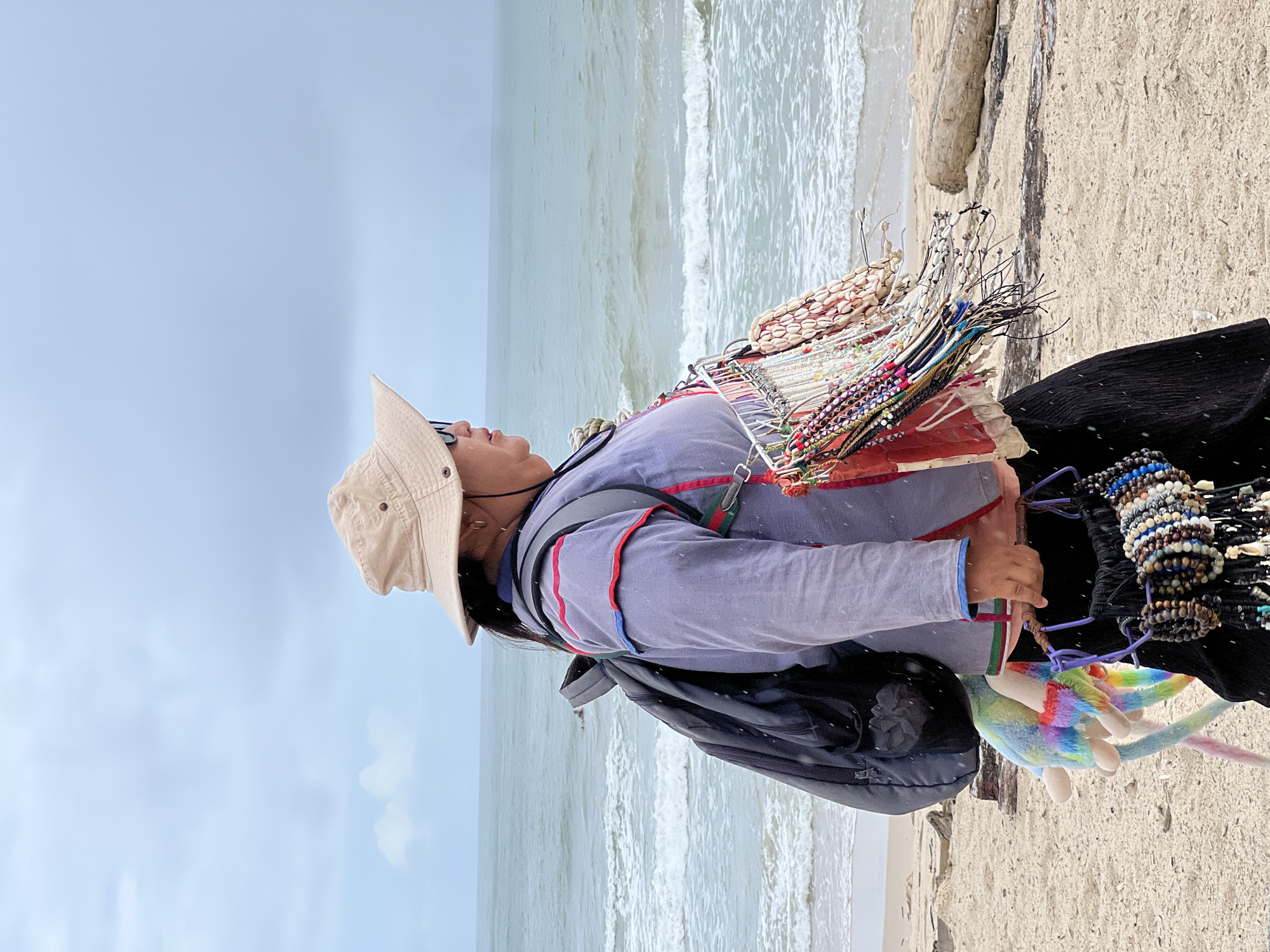 An individual wearing a wide-brimmed hat and sunglasses stands on a sandy beach, facing right, with the ocean in the background. They are carrying a backpack and have several strands of jewelry, possibly for sale, attached to their front. A colorful stuffed animal is also attached to the backpack. The sky is overcast, and there appears to be light rain. The environment is a beach with waves gently rolling onto the shore.