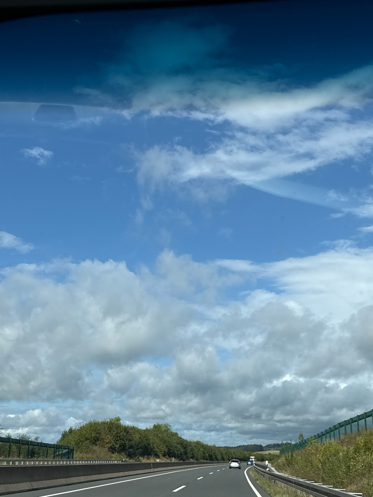 The image shows a view from inside a vehicle traveling on a highway. The sky is blue with scattered white clouds. The road ahead is paved and has white lane markings. There are vehicles on the road, including cars and a truck. On either side of the road, there are trees and vegetation. A sound barrier is visible along the right side of the highway. The image appears to be taken in Germany, possibly near Bad Sachsa.