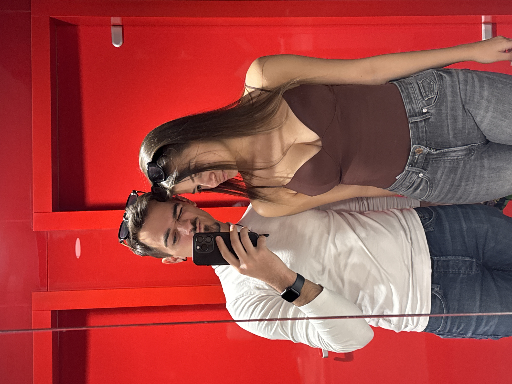 A man and a woman are taking a selfie in what appears to be a store in Sofia, Bulgaria. The background is primarily bright red. The man is wearing a white long-sleeved shirt and blue jeans, and holding a black phone. The woman is wearing a brown tank top and gray jeans. They are standing close together. The image seems to be reflected in glass or a mirror, with a slight reflection visible on the bottom. The store's interior is modern.