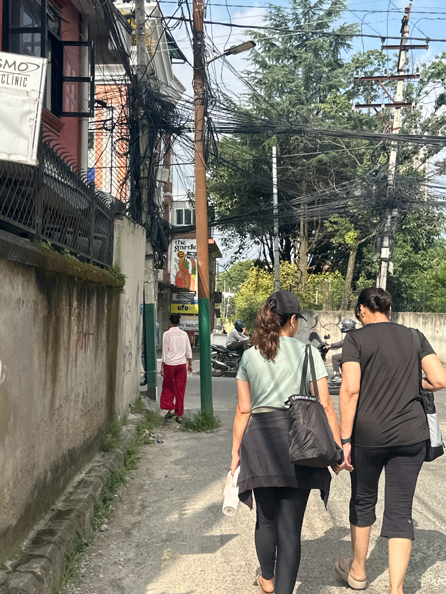 The image captures a street scene in Lalitpur, Nepal. In the foreground, two women walk away from the camera, holding hands. The woman on the left wears a light green t-shirt, a black skirt, and black leggings, carrying a white water bottle and a black bag. The woman on the right wears a black t-shirt and black cropped leggings, carrying a black and white bag. Both are wearing sandals. Further down the sidewalk, a person in a white shirt and red pants walks away from the camera, also moving away from the viewer. In the background, a building with red and white elements is visible, adorned with numerous tangled electrical wires. A sign for "the garden" and "UFO" is also present on the building. Motorcycles are parked on the side of the street. The environment appears to be an urban street with concrete sidewalks and a wall along one side. Trees and electrical poles with an extensive network of wires are prominent features of the background. The scene is illuminated by daylight, suggesting it is daytime.