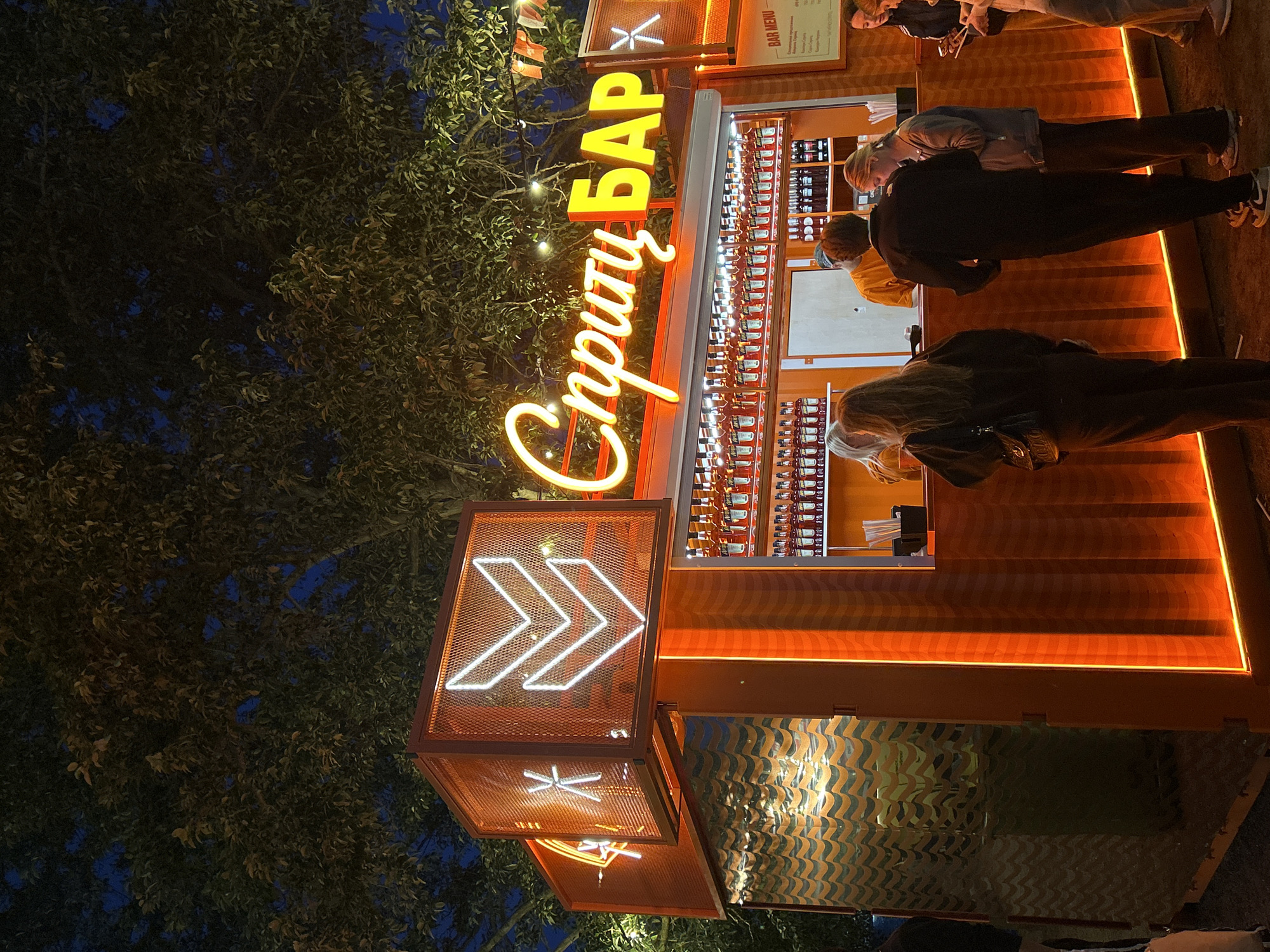 The image depicts an outdoor bar, identified by the neon sign "Спирту Бар" which translates to "Spirit Bar" in Russian. The bar is illuminated with orange neon lights and has a prominent display of numerous bottles, likely alcoholic beverages, on shelves behind the counter. Several people are visible, some standing in line to order at the bar, while others are in the process of being served. The bar itself is constructed from an orange, corrugated material and features a metallic, wave-patterned side. The scene is set at night, with trees visible in the background, suggesting an urban outdoor setting. A bar menu is also visible to the right of the main sign, though the text is largely illegible. The overall ambiance is lively and festive, likely a popular spot for evening entertainment.