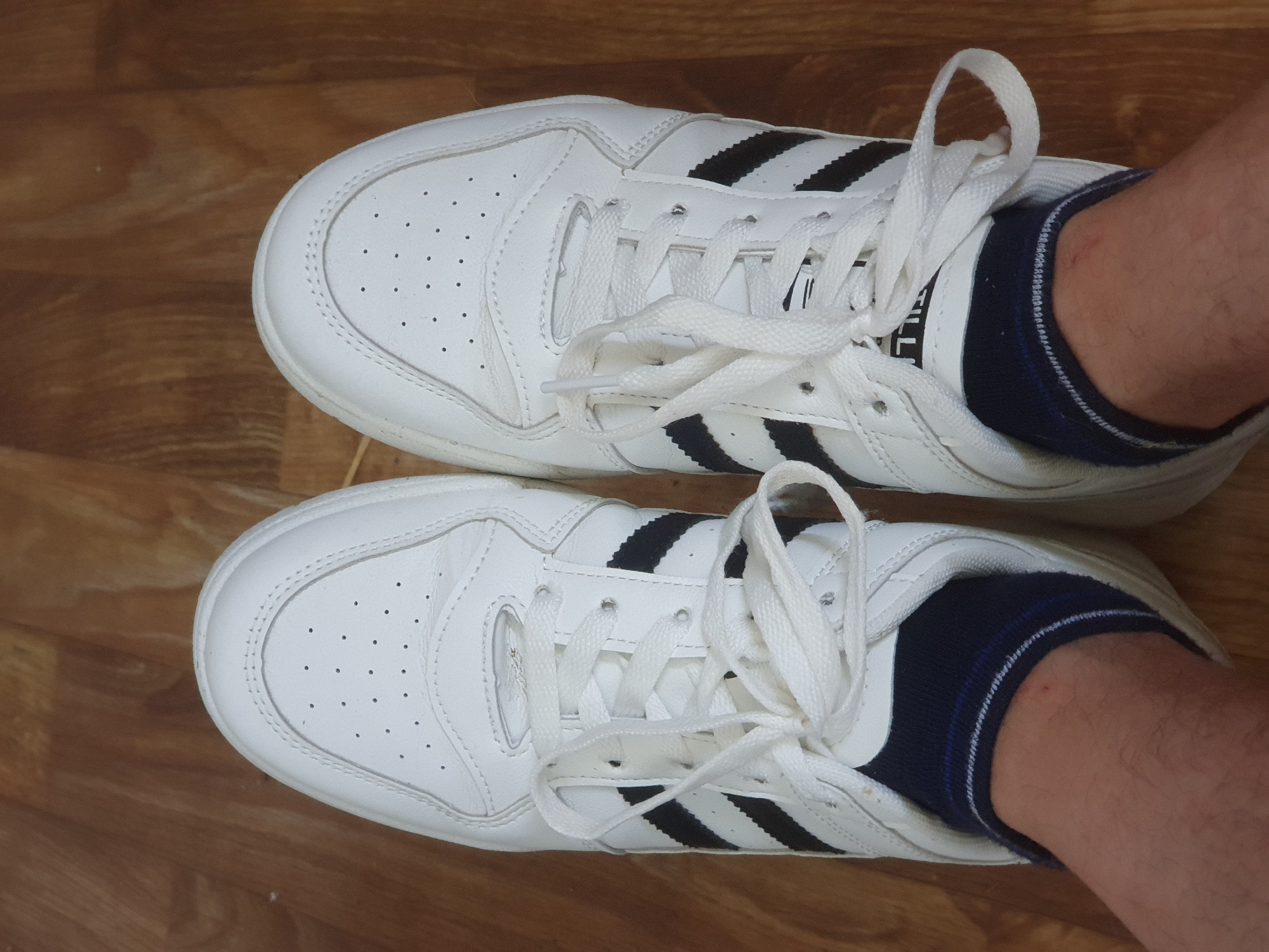 A pair of white Adidas sneakers with black stripes and white laces are shown from a top-down perspective. The sneakers are worn by an individual whose lower legs are visible, covered by dark socks with blue and white stripes. The scene takes place indoors on a wooden floor. There is no visible indication of the location being in Zubrești, Moldova.