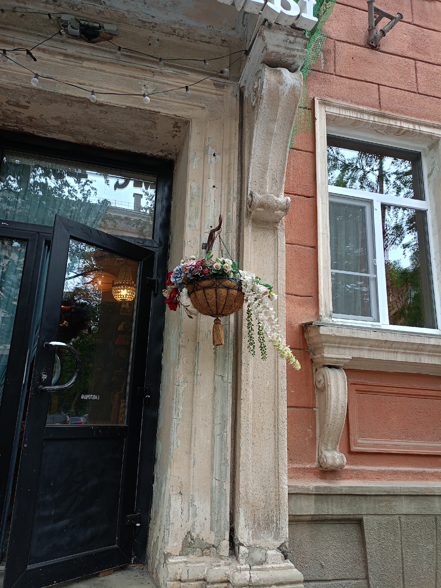 The image shows the exterior of a building in an urban setting, likely a storefront. A dark-colored door is partially open, revealing a glimpse of the interior which features a hanging chandelier. Adjacent to the door, a basket filled with artificial flowers is suspended from a hook. The building's facade is made of brick, painted in an orange-red hue, with decorative architectural elements. A window with white framing and a white mesh screen is visible on the right side of the image. The wall above the door is weathered and shows signs of peeling paint. A string of bare light bulbs is mounted along the top of the entryway. The ground in front of the door appears to be paved. The overall impression is one of an aged building with some decorative touches.
