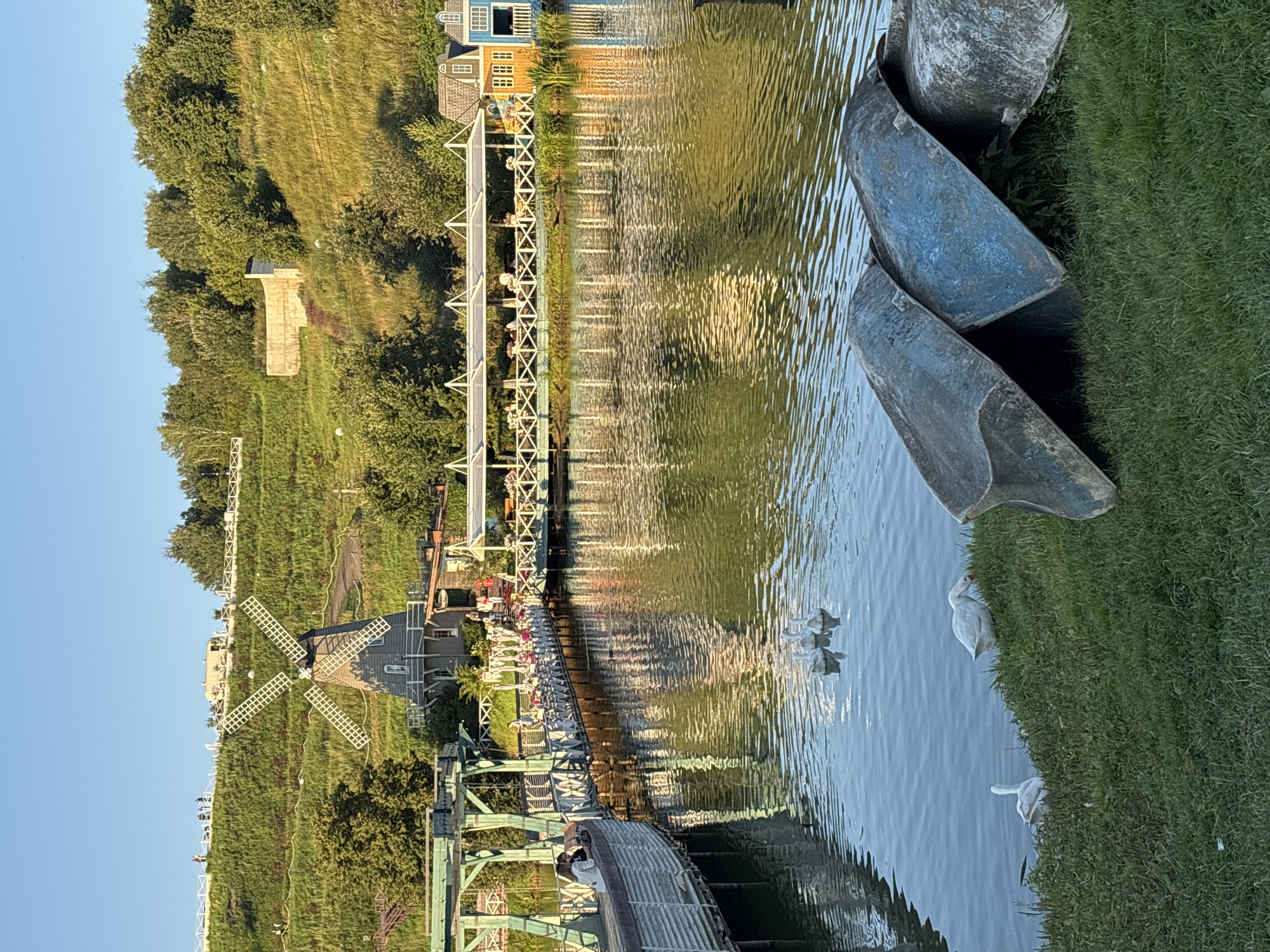 The image depicts a picturesque scene at a park in Romashkovo, Russia. In the background, a lush green hillside is dotted with trees and a white fence, with a traditional Dutch windmill situated on its slope. A prominent wooden bridge, painted white with green accents, spans a tranquil lake. Several people are seen walking on and around the bridge, and a covered seating area with tables and chairs is set up along its length, with some patrons seated. The lake reflects the surrounding greenery and structures. In the foreground, several white geese are visible on the grassy bank and in the water. A stack of overturned boats lies on the grass to the right of the frame. The sky is clear and blue, indicating a sunny day.