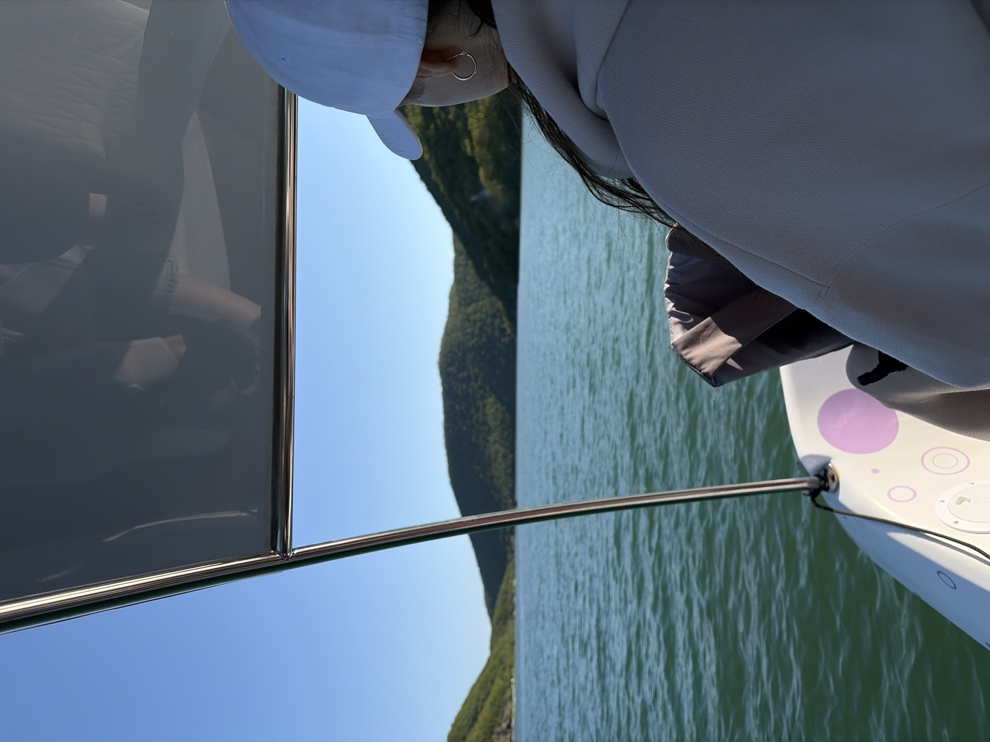 A person in a white cap and light-colored clothing stands on a boat on the water, looking out towards a green, tree-covered shoreline under a clear blue sky. The person's left ear is visible, showing a simple hoop earring. The boat features a metal railing and a window-like panel on the left side, and a portion of the boat's hull, decorated with purple circles, is visible on the right. The location is likely near Abrau-Dyurso, Russia, based on the type of scenery (water and hillside vegetation).