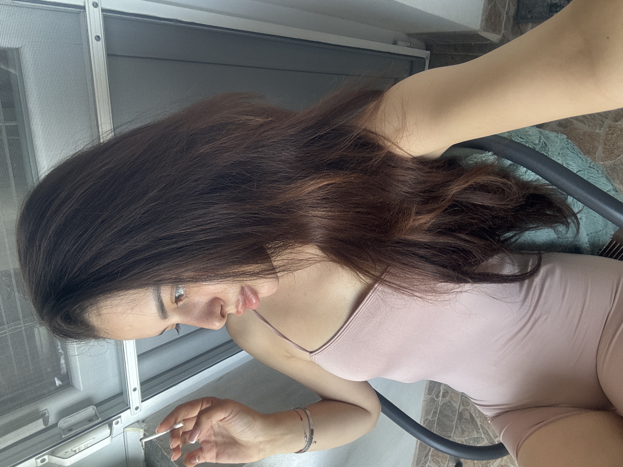 A woman is depicted in a profile view, looking downwards. She has long, wavy, dark brown hair that cascades over her shoulder and back. She is wearing a light pink or nude-colored, form-fitting tank top or bodysuit. Her right arm is visible, featuring a thin bracelet on her wrist, and her fingers, adorned with manicured nails (possibly French tip), delicately hold a slim, white, cylindrical object, resembling a cigarette or vape pen.
She appears to be seated on a chair, with a portion of its black frame and a light blue or teal furry blanket/cushion visible. The background shows a white metal frame, likely part of a window or screen door with a mesh screen, suggesting an indoor or balcony setting. The floor beneath her features light-colored tiles. The lighting indicates it is daytime. Her expression is neutral and focused on the object in her hand or something below. There is no visible text in the image. The scene is set in Plovdiv, Bulgaria.