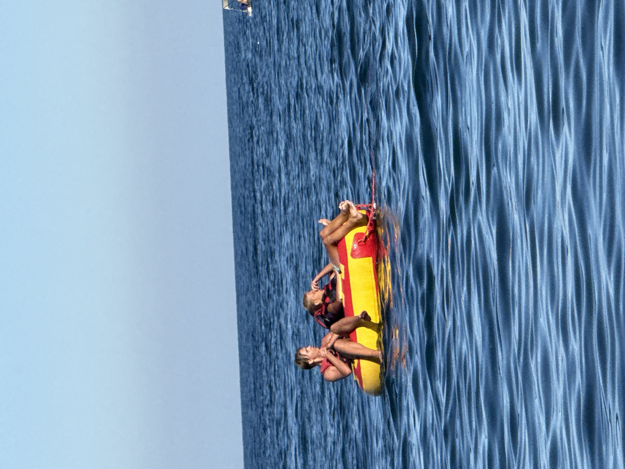 Two individuals are seated in a yellow and red inflatable tube on the surface of a blue body of water. The person on the left is a male with blonde hair, wearing a red life jacket, and is looking to his left. The person on the right is a female with blonde hair, also wearing a red life jacket, and appears to be resting her head on her hand, looking to her right. The water is calm with gentle ripples. The sky is clear and a light blue. In the far distance, a white structure, possibly a boat or a jet ski, is visible on the water. The location is the Black Sea near Partenit, Ukraine.