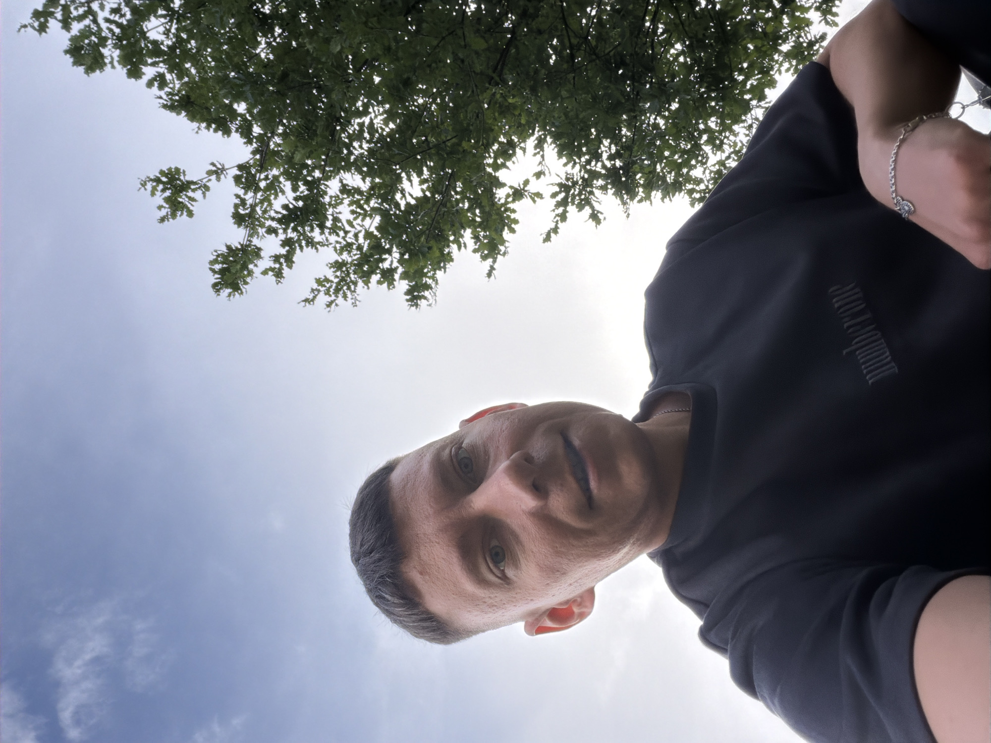 A low-angle, wide shot captures a man in his late 20s or early 30s, looking up at the camera. He has a short, buzzed haircut with a fade and light-colored eyes. The man is wearing a dark t-shirt with a subtle embroidered logo on the left chest that reads "numberone". He is also wearing a silver bracelet on his left wrist.
The background consists of a clear blue sky with some wispy clouds, and a large, leafy green tree branches out to the right of the frame. The lighting suggests it is daytime, possibly late afternoon or early evening due to the soft light.
The image does not contain any specific information about its location in Moscow, Russia.