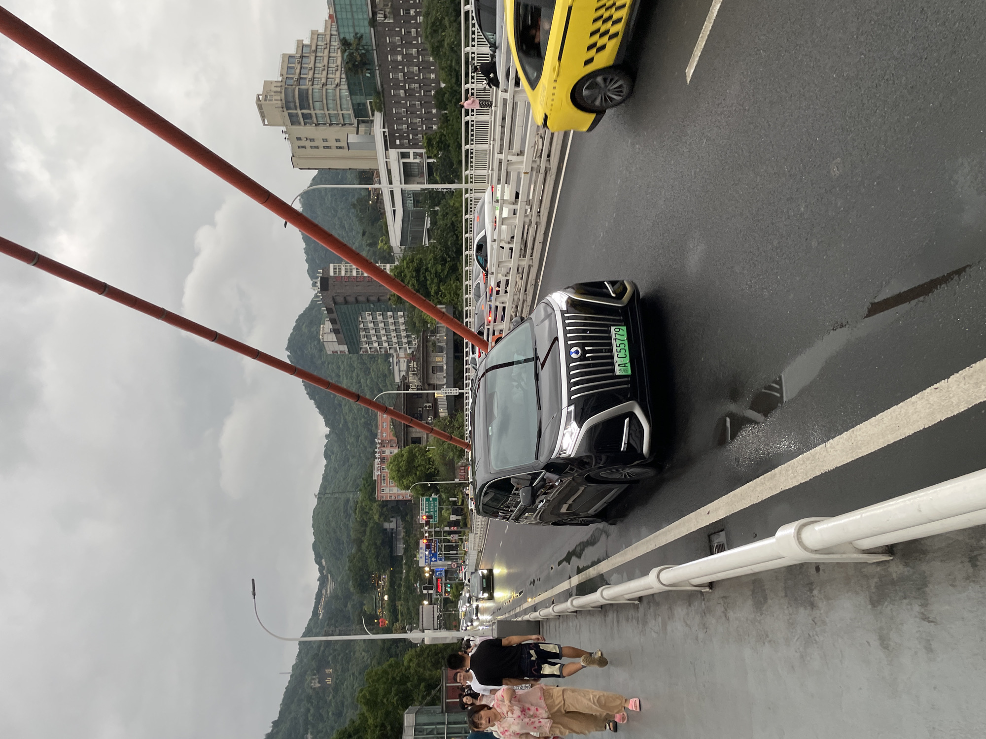 The image depicts a street scene on a bridge in Nan'an District, China, under an overcast sky. On the left side of the frame, a pedestrian walkway features several people walking. Among them is a woman in a light-colored floral top and beige pants, and a man in a black t-shirt and patterned shorts, both walking away from the camera. A white railing separates the walkway from the road.
The road itself is wet, suggesting recent rain, with visible puddles. Several vehicles are present. In the foreground, a black luxury van with a distinctive grille and a green license plate reading "A C55779" is prominently featured. To its right, a yellow taxi with a black checkered pattern on its side is also visible. Further down the road, other cars and vehicles are driving or stopped in traffic.
In the background, lush green mountains rise, dotted with buildings of varying architectural styles, including apartment complexes and what appear to be commercial structures. Red cables, part of the bridge's suspension system, are angled across the upper portion of the image, contrasting with the muted tones of the sky and landscape. Streetlights and traffic signals are also visible along the bridge. The overall atmosphere suggests a busy urban environment during daylight hours on a cloudy day.