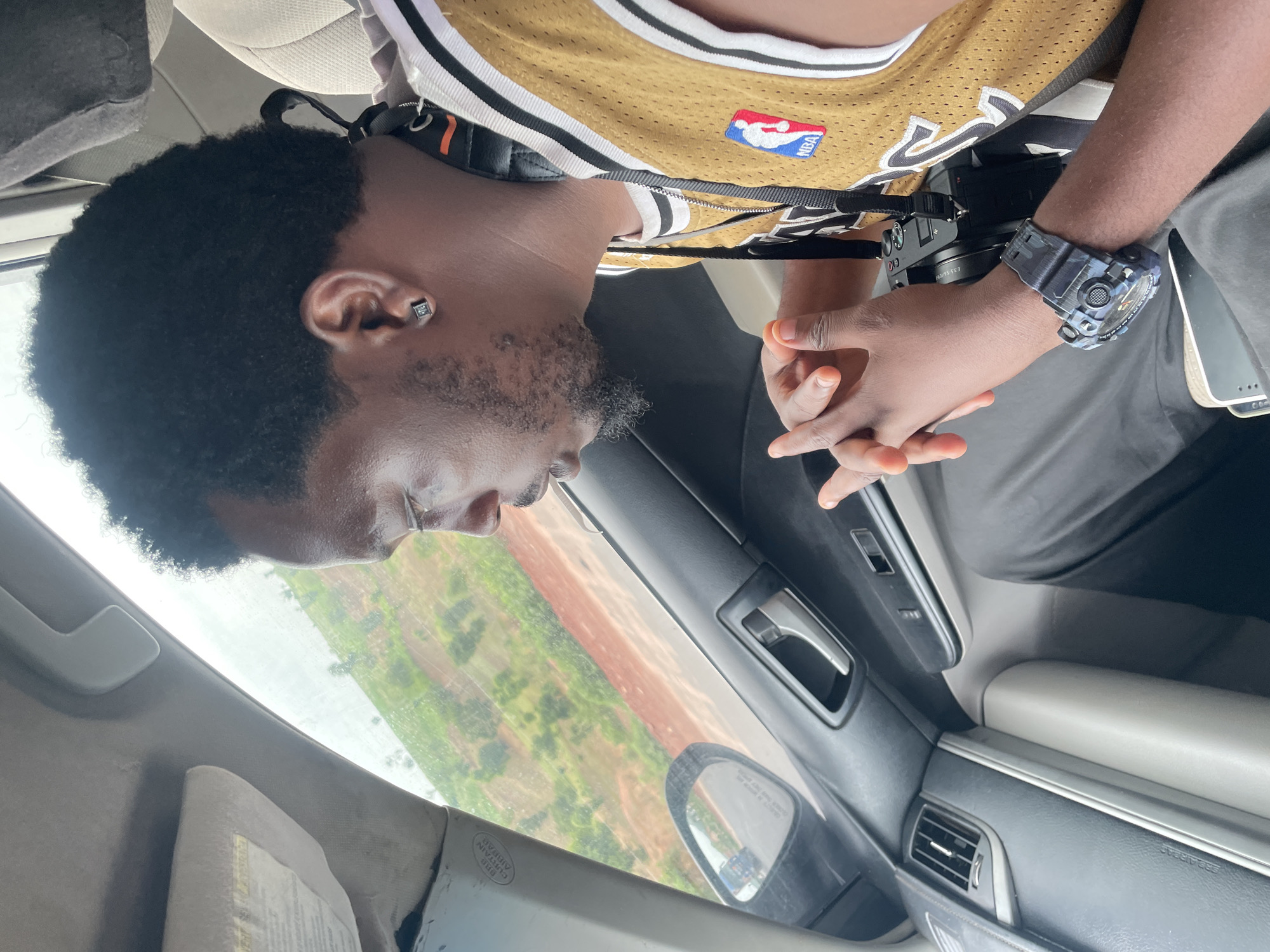 The media file shows a person sitting in the passenger seat of a car, looking out the window. The person is wearing a gold NBA jersey and has a camera around their neck. They are also wearing a camouflage wristwatch on their left wrist. The car appears to be in motion, as evidenced by the blurred scenery outside the window. The scenery outside suggests a rural or semi-rural location, with greenery and red soil visible. The lighting suggests it is daytime. The person's expression is neutral. The image is shot from a slightly high angle, looking down at the person.
