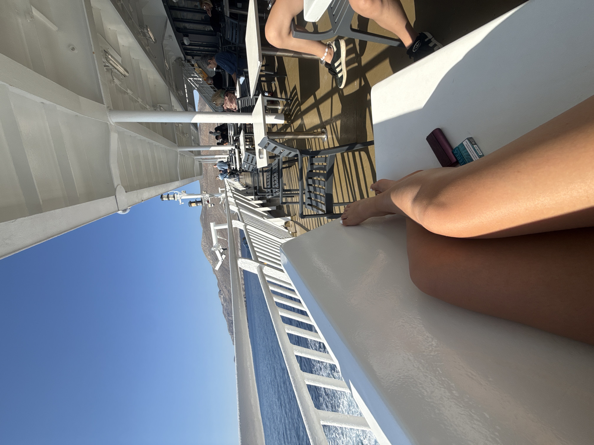 A first-person perspective shot from a ferry, looking forward and to the right. The foreground shows a person's tanned legs resting on a white surface, with a pack of HEETS tobacco sticks and a dark purple wallet on a white table. The sea is visible to the left, with white railings and a white structure of the ferry. Further into the scene, other passengers are seated at tables and chairs under a white awning, with a view of a mountainous coastline in the distance under a clear blue sky. The image was taken in the city of Livadia, Greece, likely on a ferry traveling along the coast.