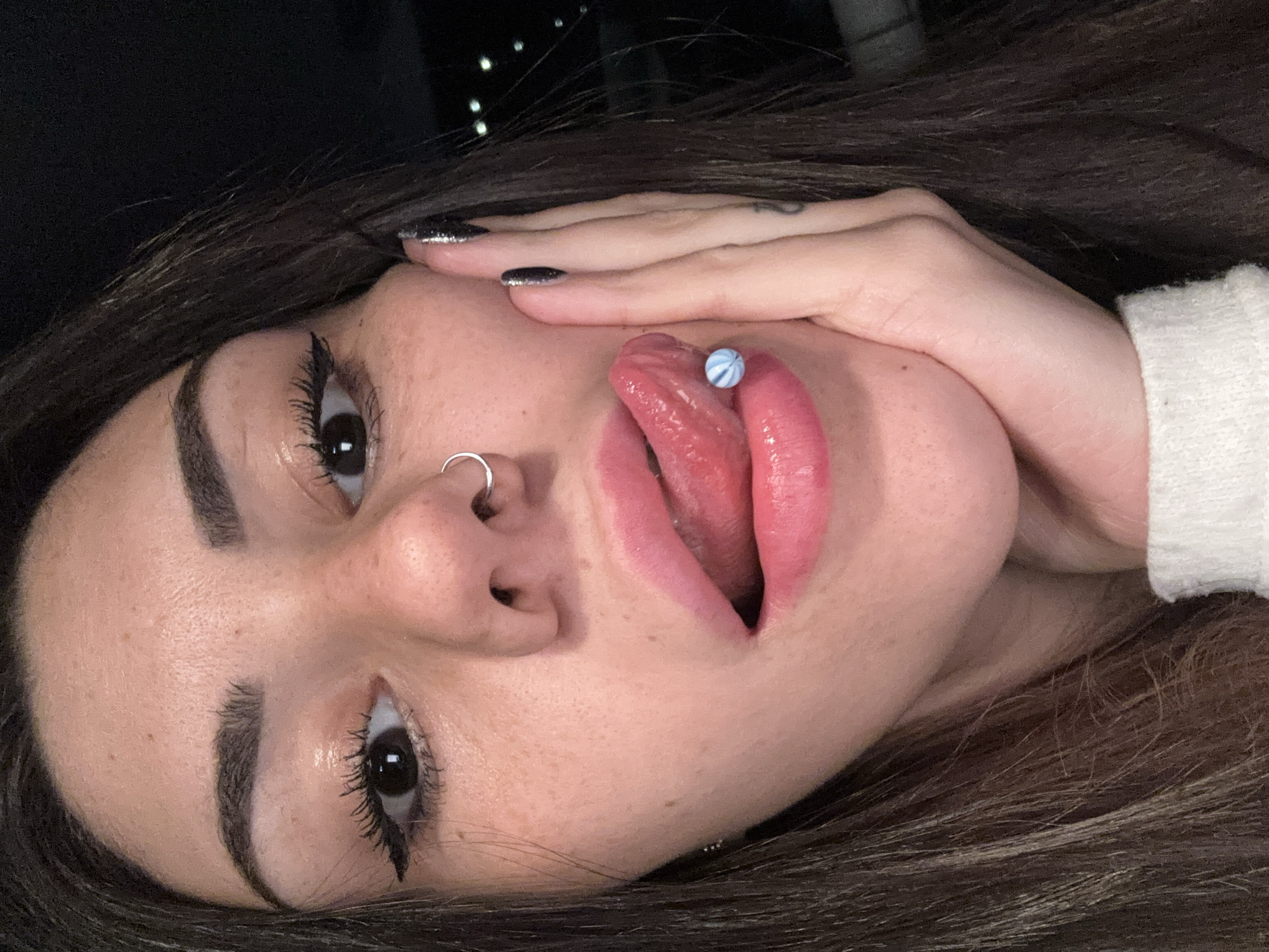 A close-up, portrait-style image of a young woman with light skin and dark hair. She is lying on her side, angled upwards towards the camera. Her mouth is open with her tongue extended, displaying a tongue piercing with a blue and white bead. She has a nose ring in her left nostril. Her left hand is positioned near her face, with silver glitter nail polish on her fingernails. Her eyes are dark, and she wears eyeliner and mascara. The background is dark with blurry lights, but the city of Sofia, Bulgaria cannot be determined in the image.